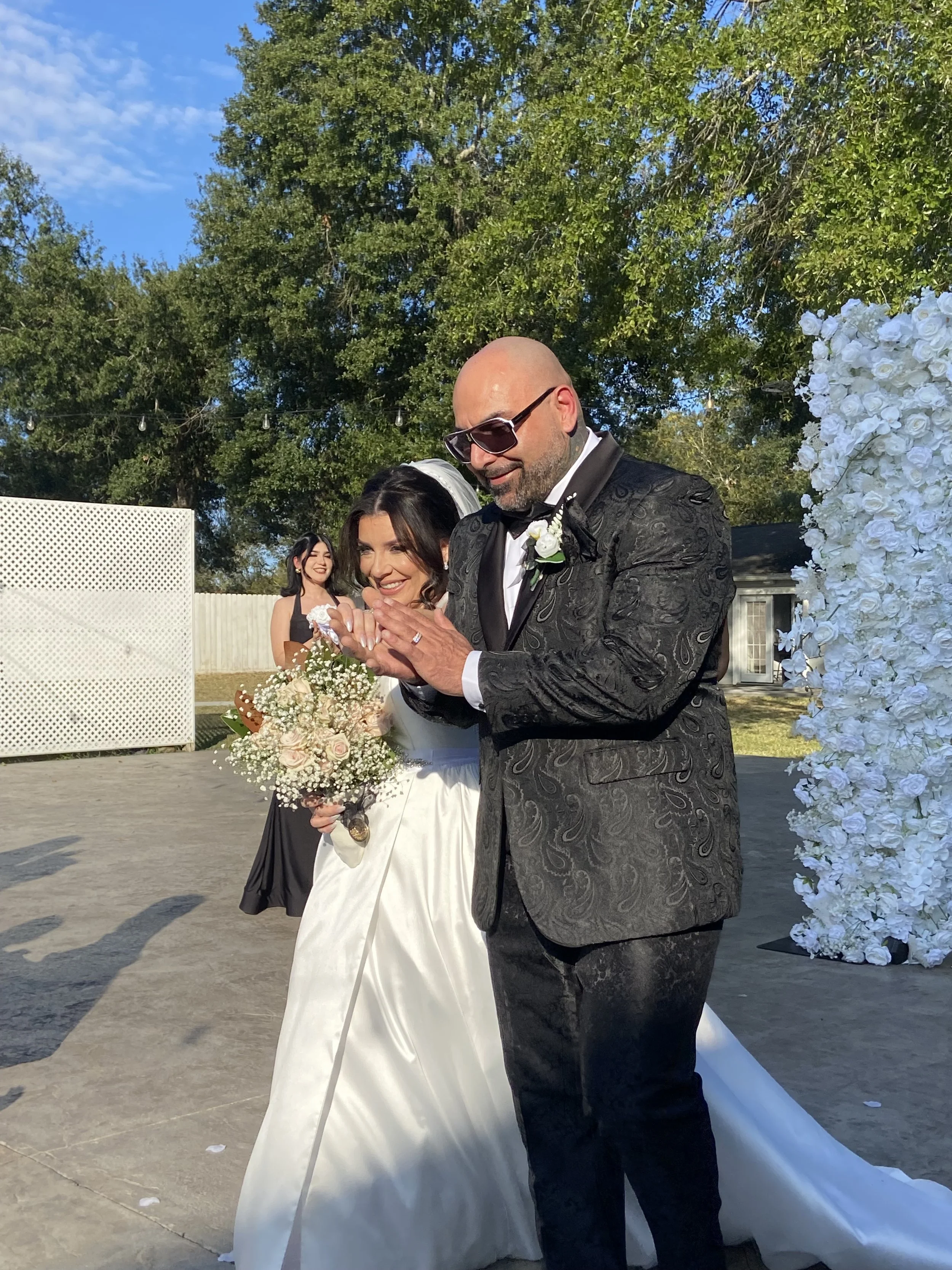 Bride and groom celebrating outdoors at their wedding, with the bride holding a bouquet of flowers and the groom wearing sunglasses and a black tuxedo with a white boutonniere. A woman in a black dress stands behind them, smiling.