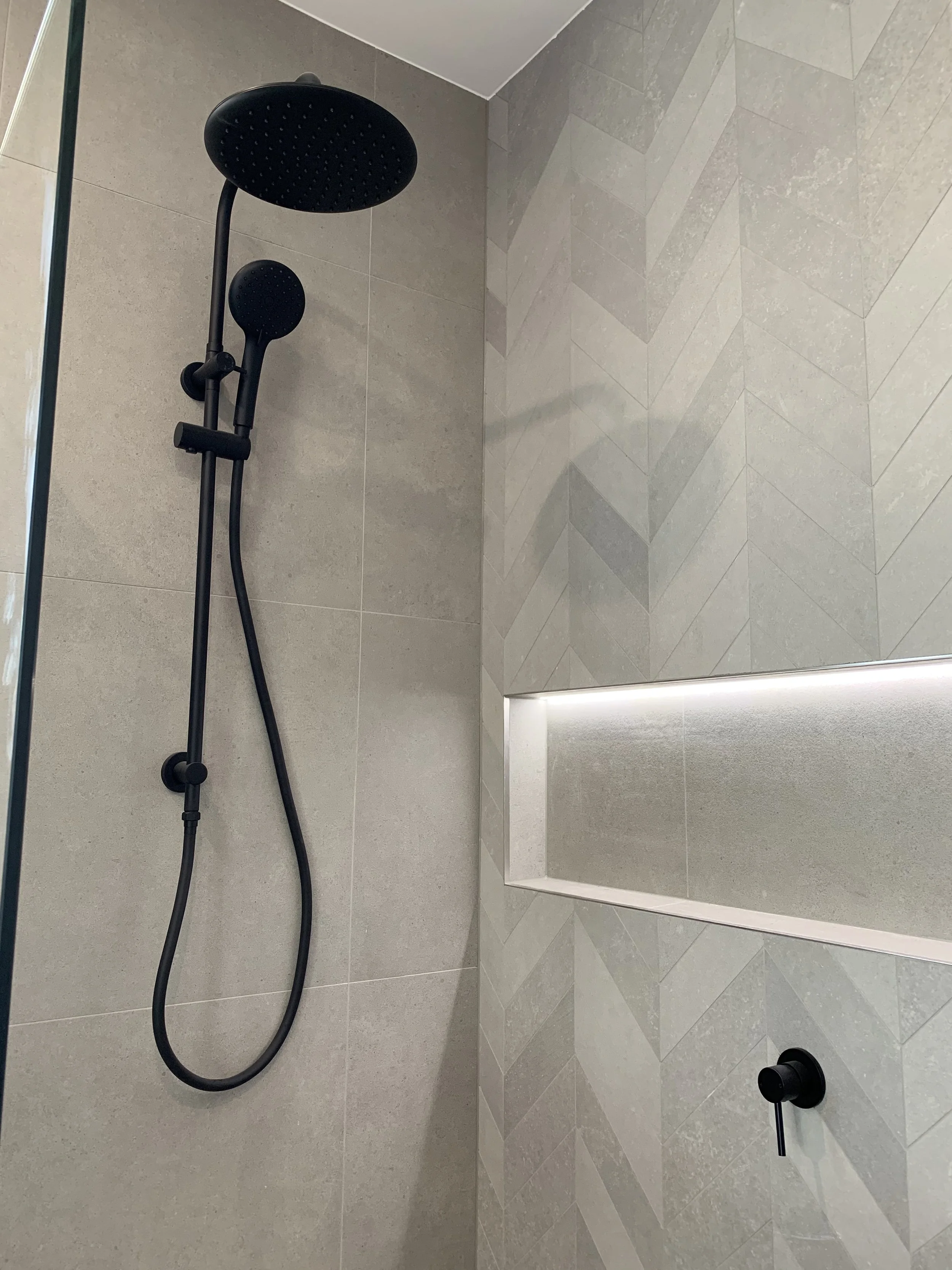 Black showerhead with an adjustable handheld shower in a modern tiled shower with a built-in shelf.