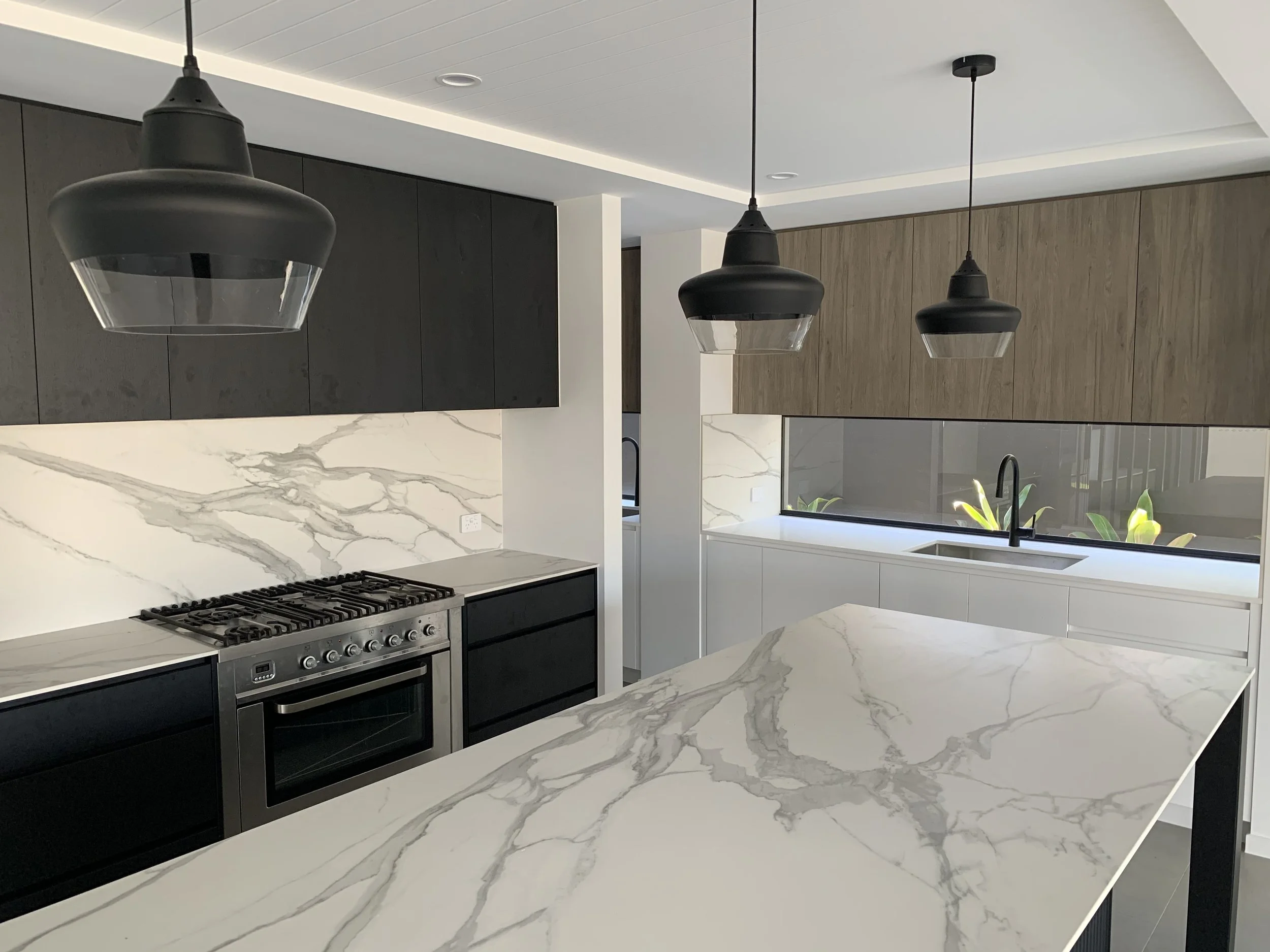 Modern kitchen with white marble countertops and backsplash, black and wooden cabinets, black pendant lights, and a window above the sink with green plants outside.