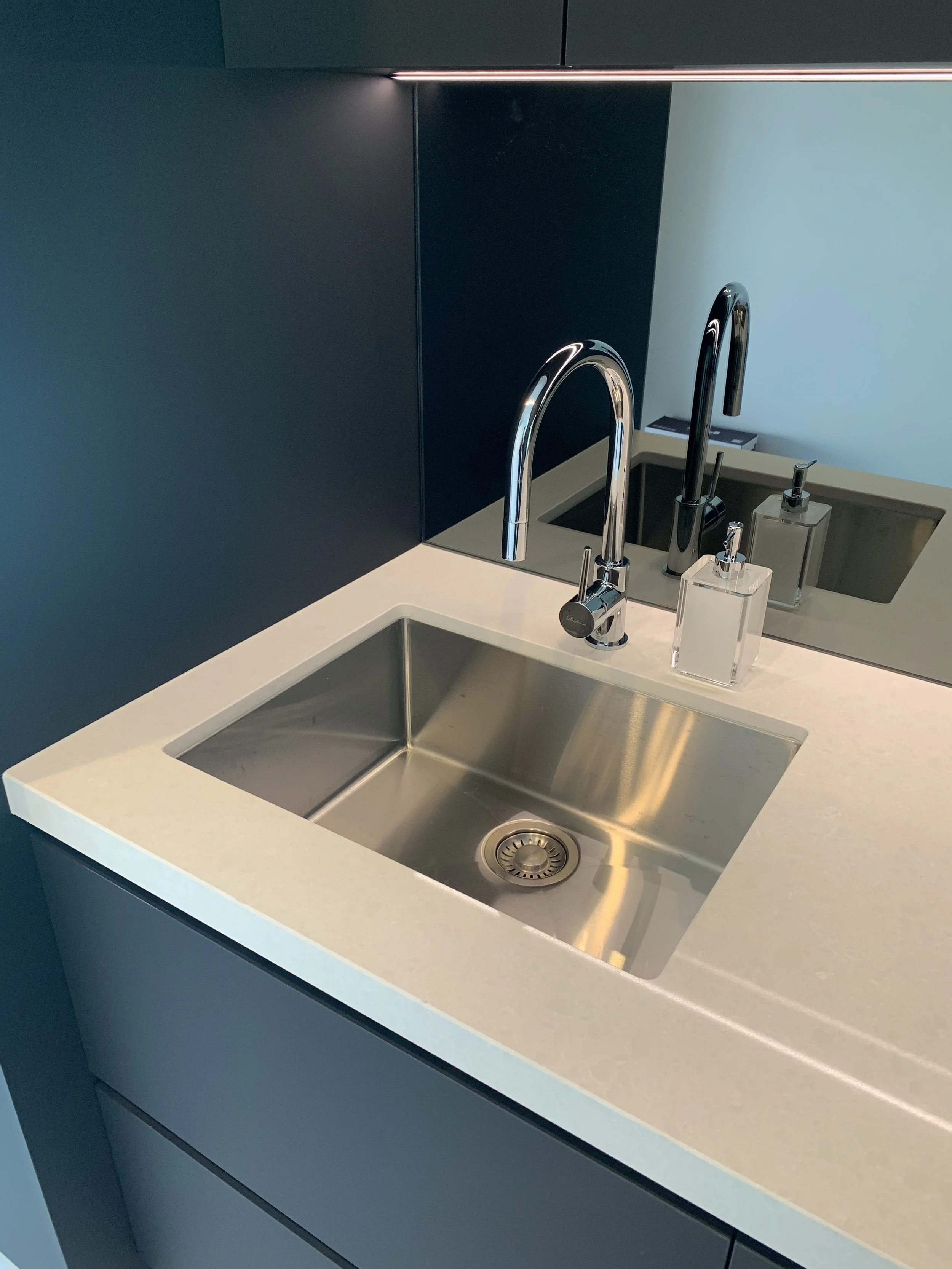 A modern kitchen sink with a stainless steel basin, chrome faucet, and soap dispenser on a white countertop, with a mirror reflecting the sink and part of the kitchen.