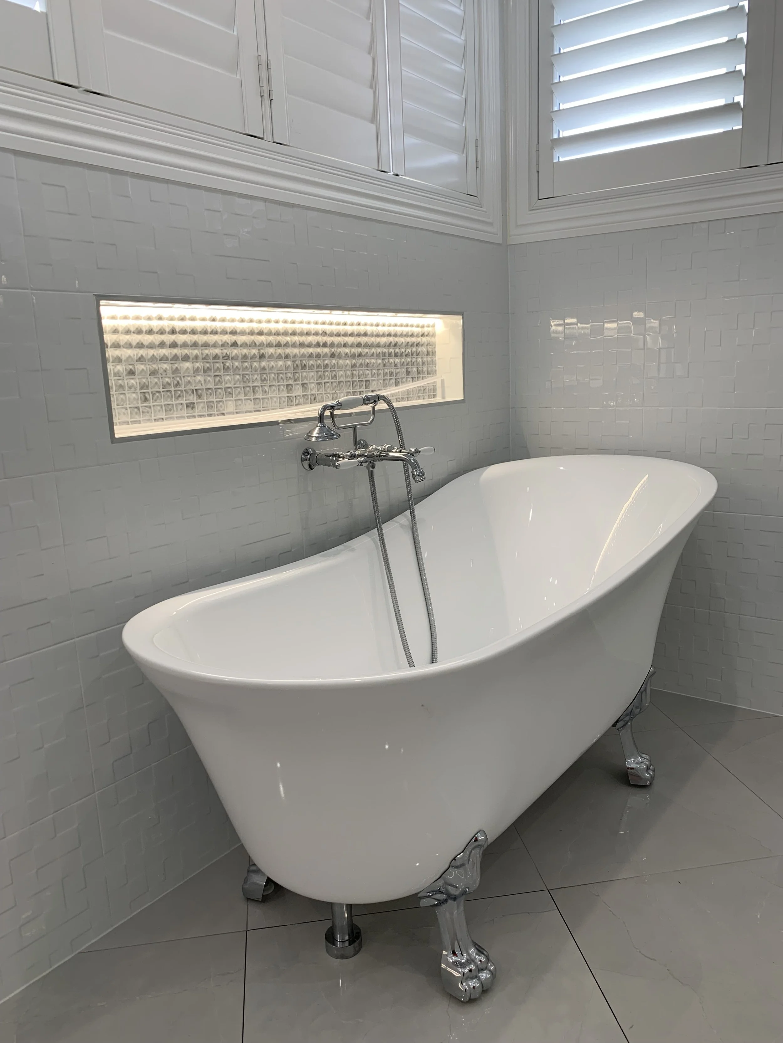 White clawfoot bathtub in a bathroom with gray tiled walls and a window with white shutters.