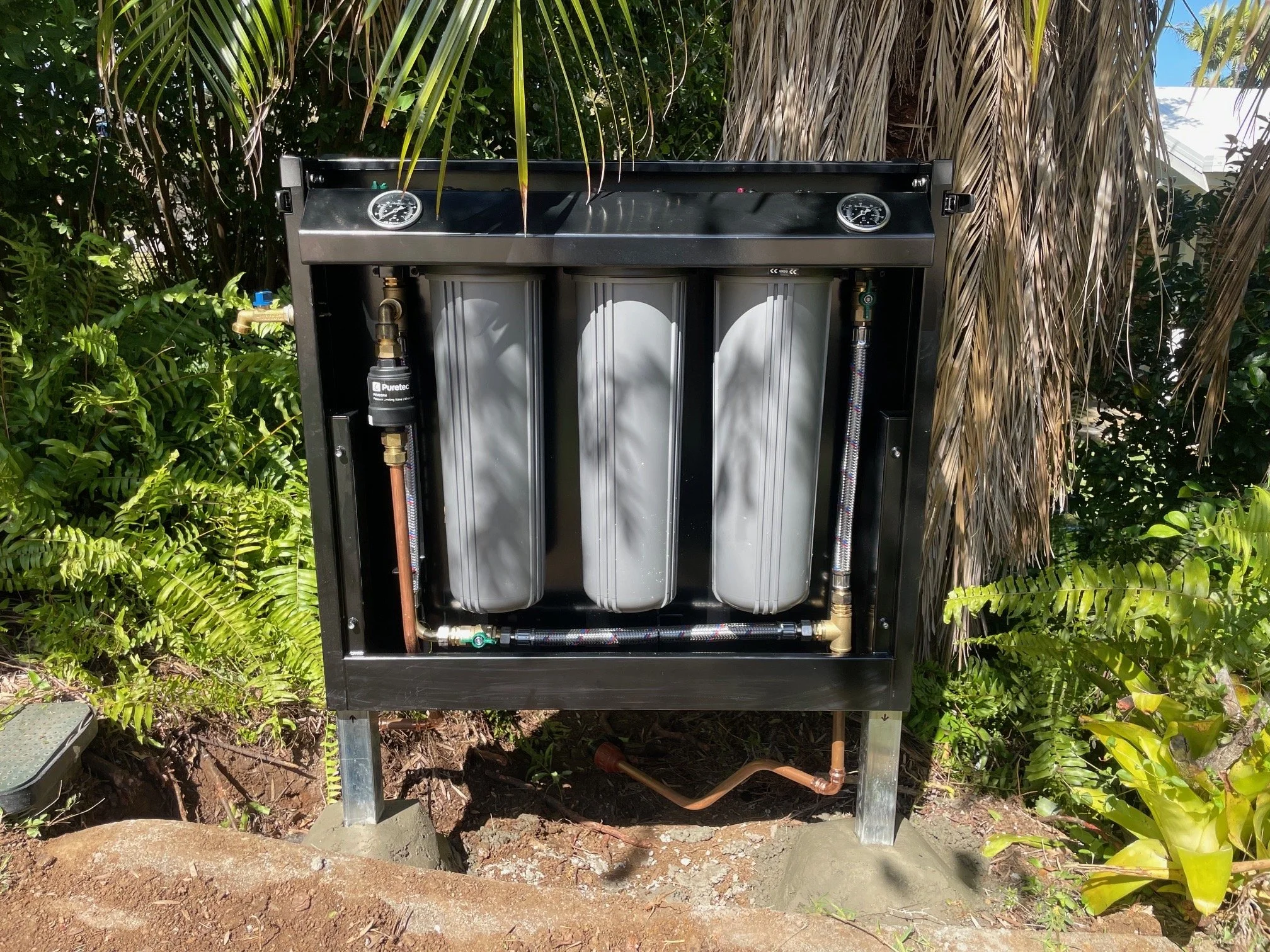 An outdoor water filtration or reverse osmosis system with three large gray cylindrical filters, connected to copper and flexible tubing, set against lush green foliage.