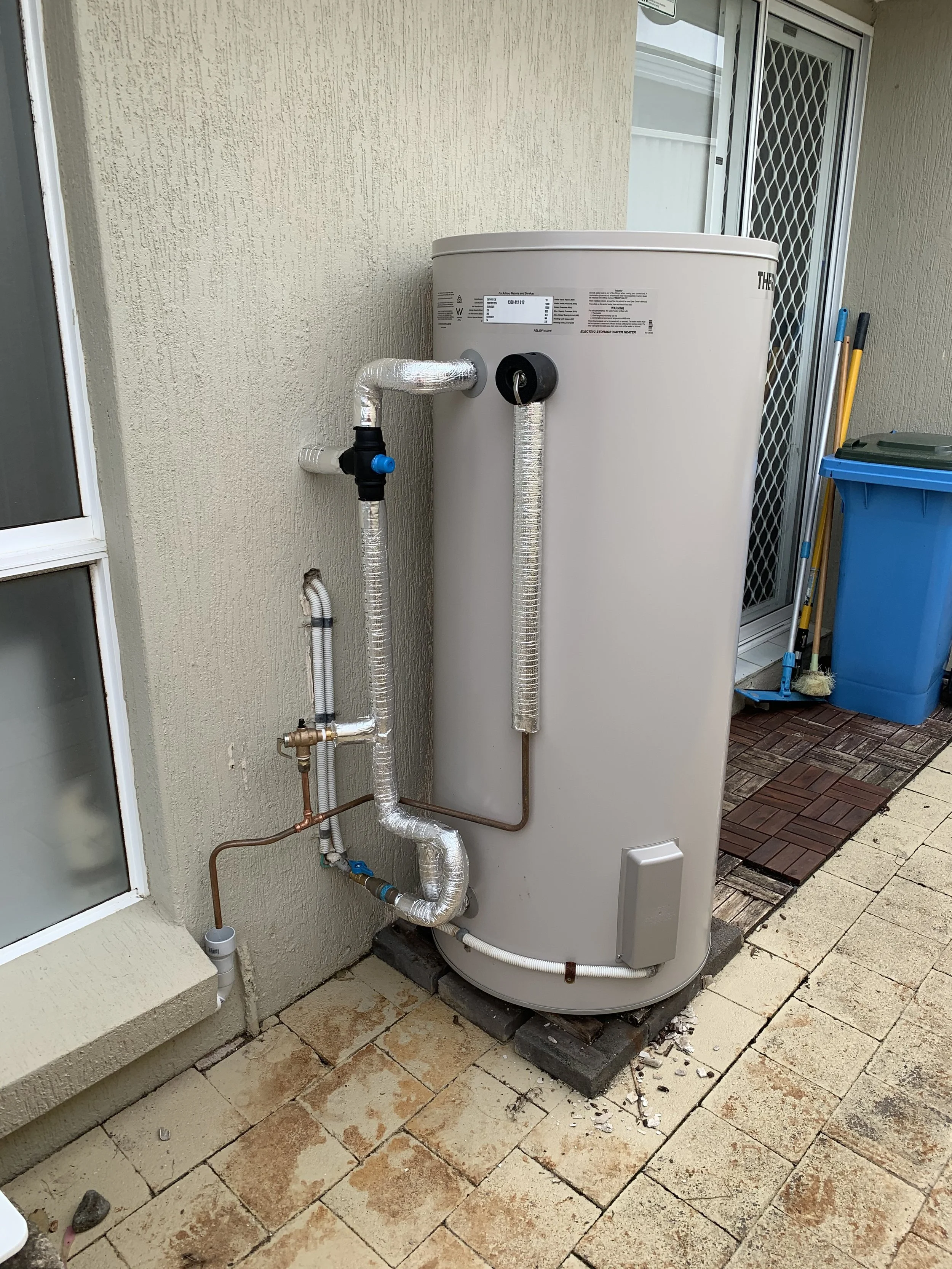 Outdoor water heater with insulated pipes, installed next to a beige wall and a sliding door, with cleaning tools and a blue trash bin nearby.