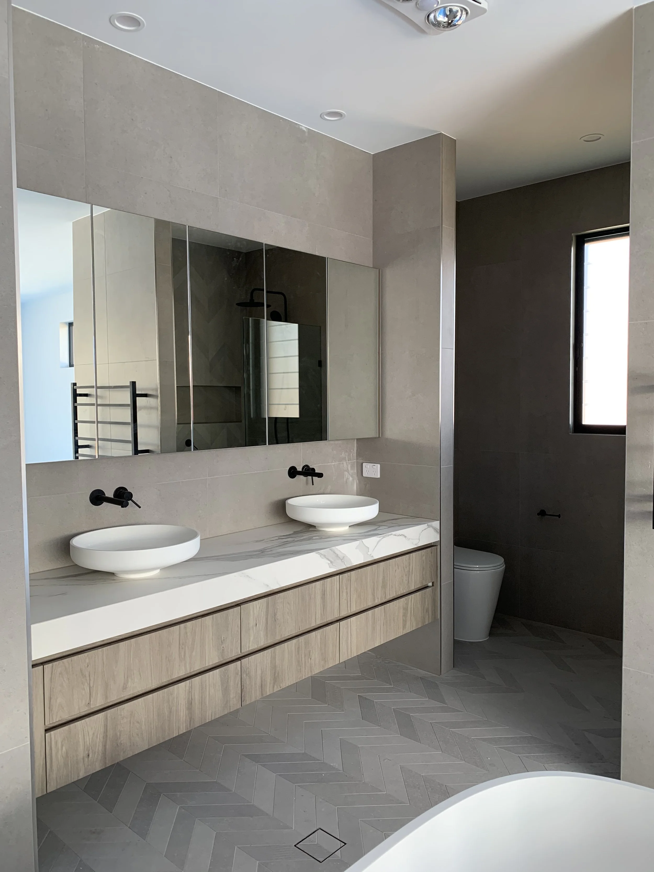 Modern bathroom with double vanity, round vessel sinks, large mirror, walk-in shower with glass door, wall-mounted faucets, and a separate toilet area with window, neutral color palette, and tiled floors.