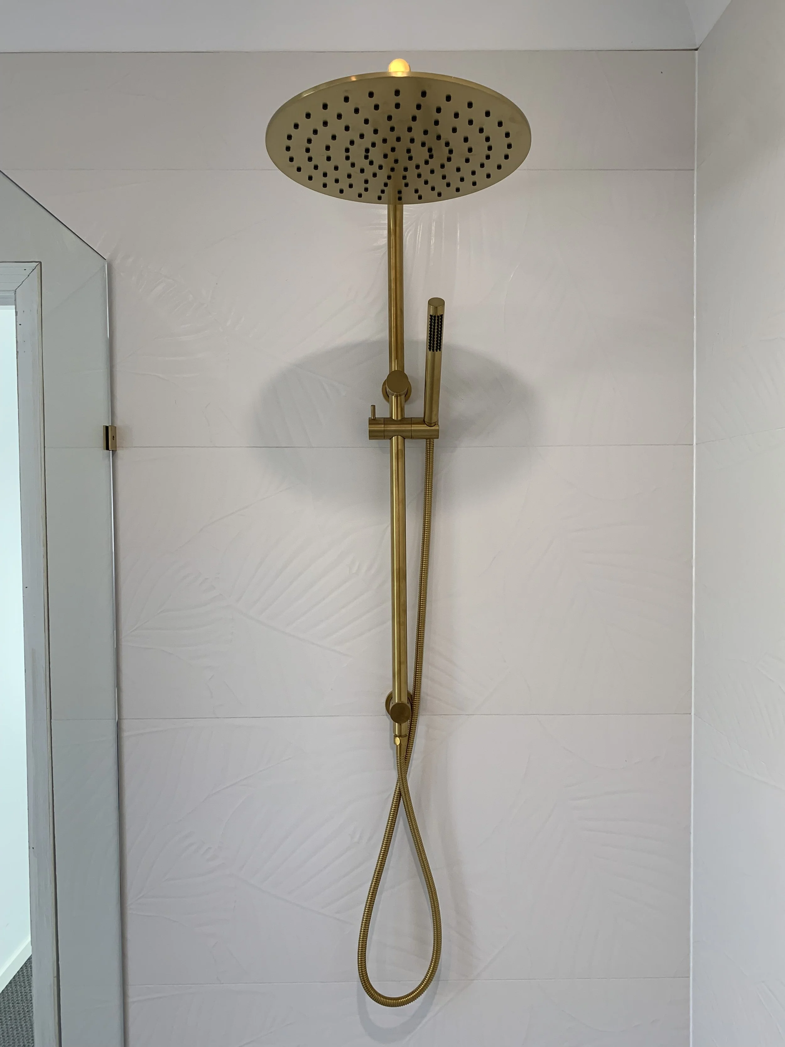 Brass rain shower with handheld sprayer mounted on a white tiled wall in a bathroom.