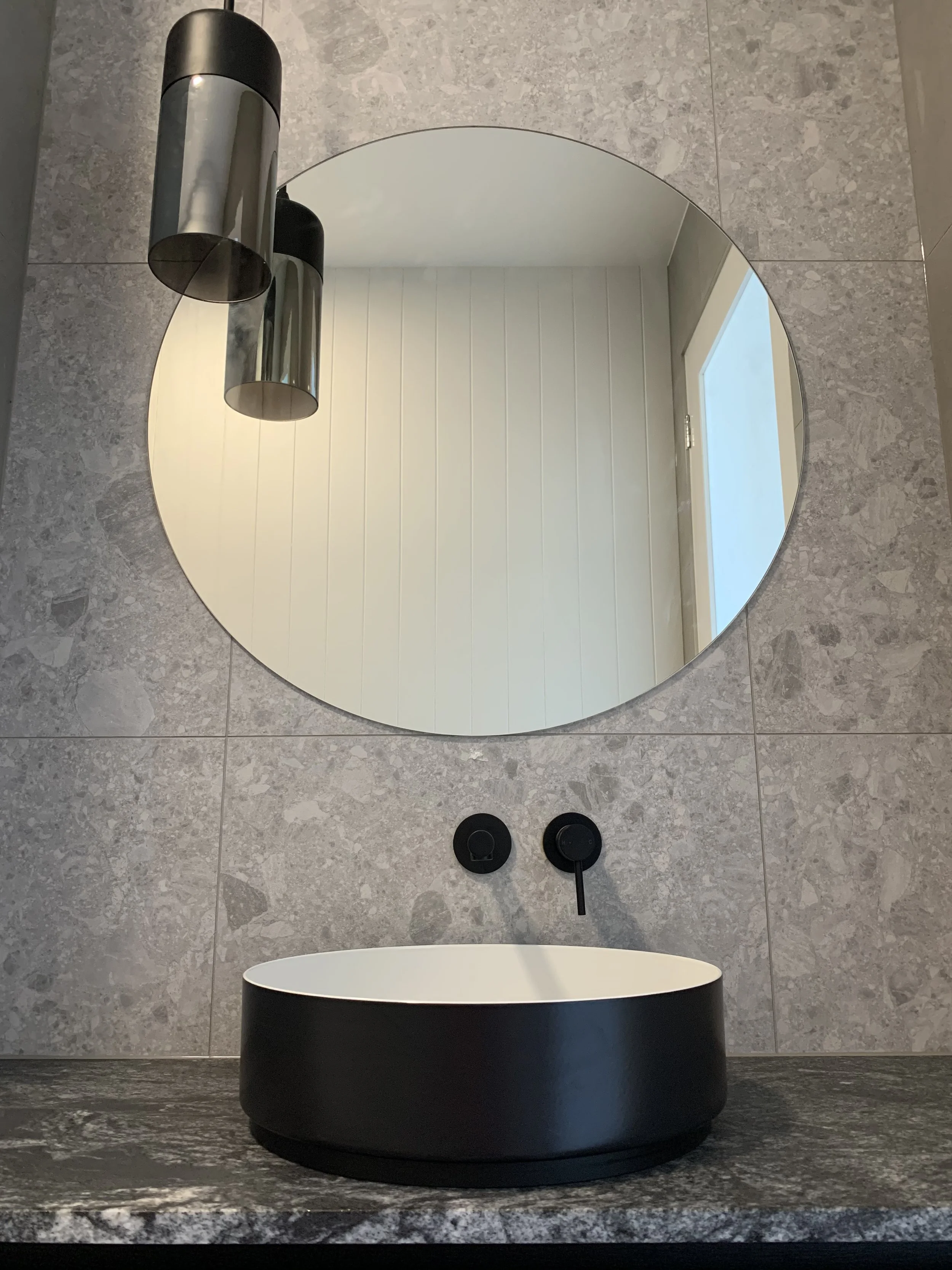 Modern bathroom sink with black exterior and white interior, round mirror on tiled wall, black faucet and controls, with windows reflected in the mirror.