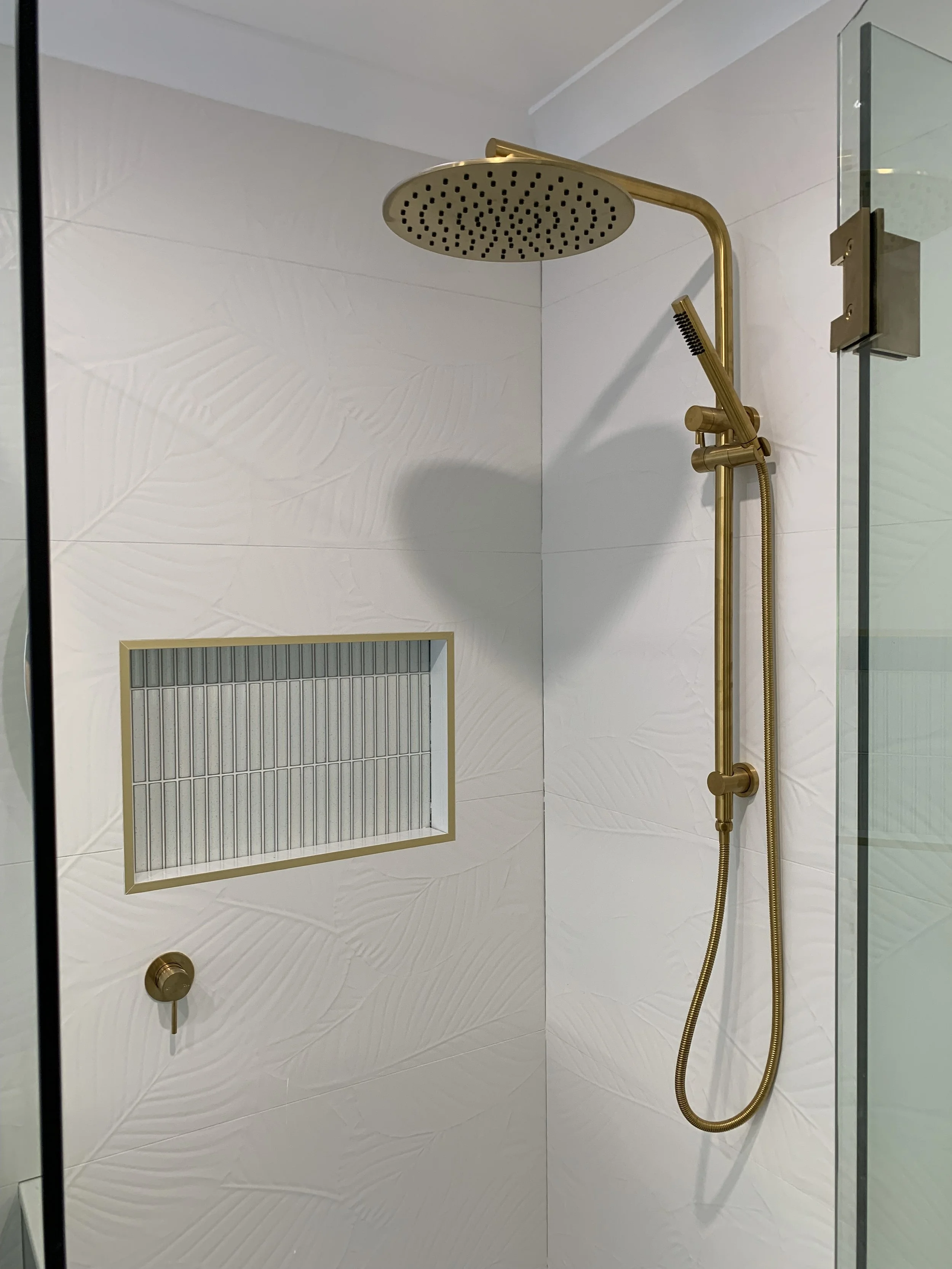 Modern bathroom shower with a gold rainfall showerhead and handheld shower attachment, white textured wall tiles, a built-in tile shelf, and a glass shower door.