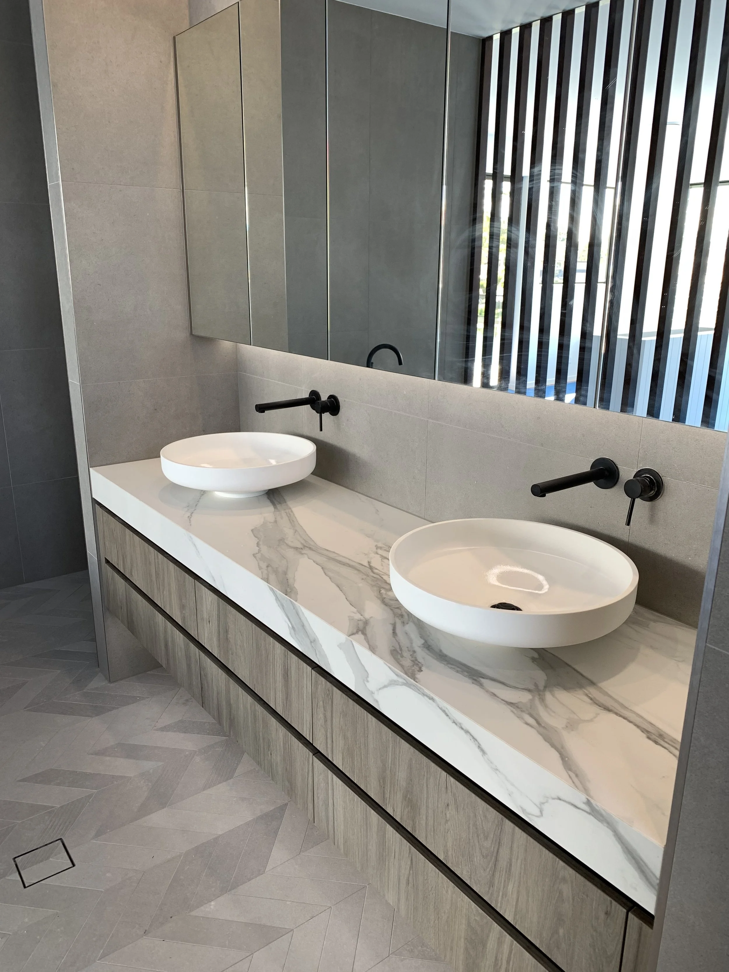 Modern bathroom with double white vessel sinks on a marble countertop, black wall-mounted faucets, a large mirror, and gray tiled walls and floors.