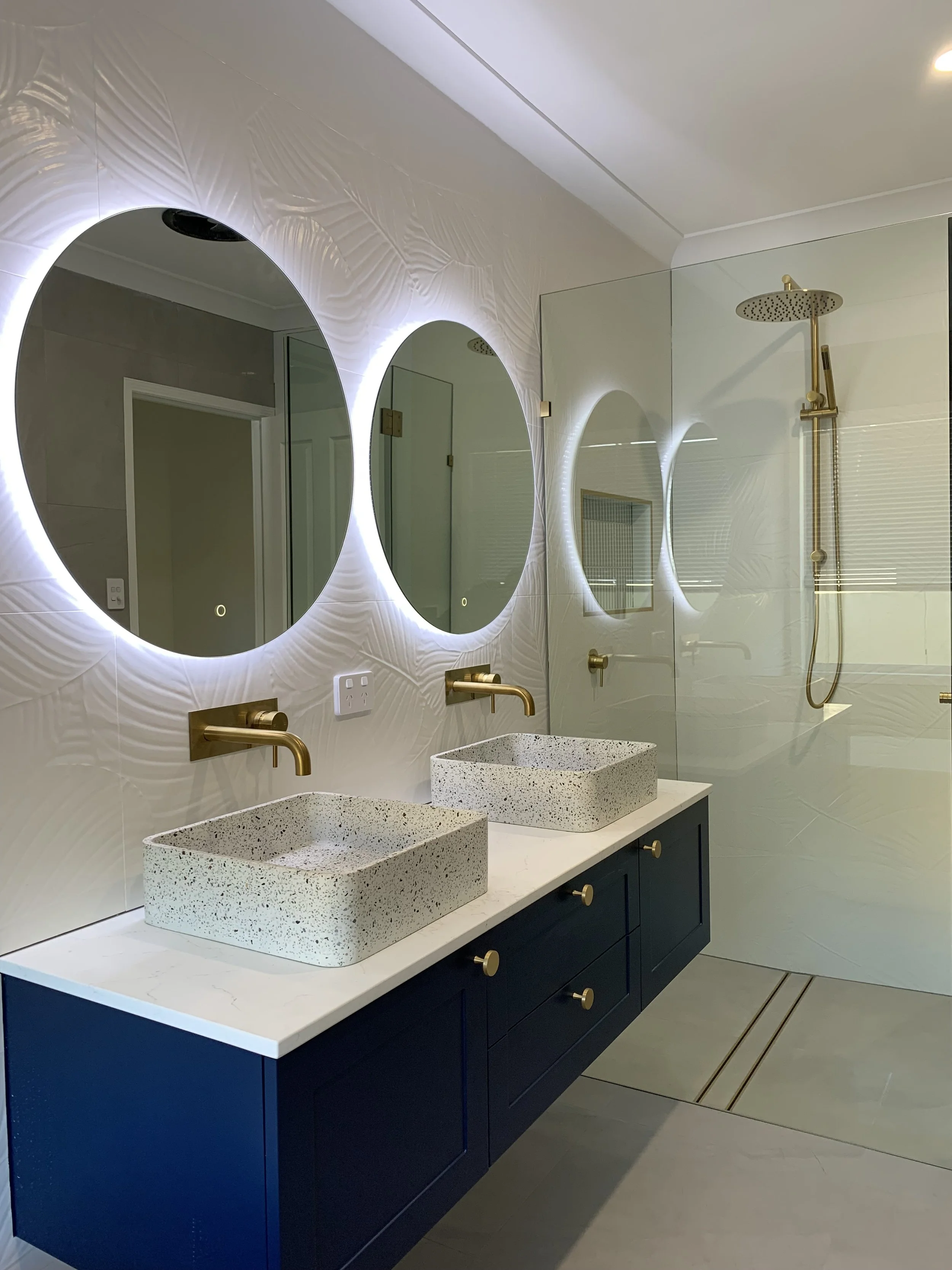 Modern bathroom with two terrazzo vessel sinks, gold fixtures, backlit mirrors, and a glass-enclosed walk-in shower with gold showerhead.