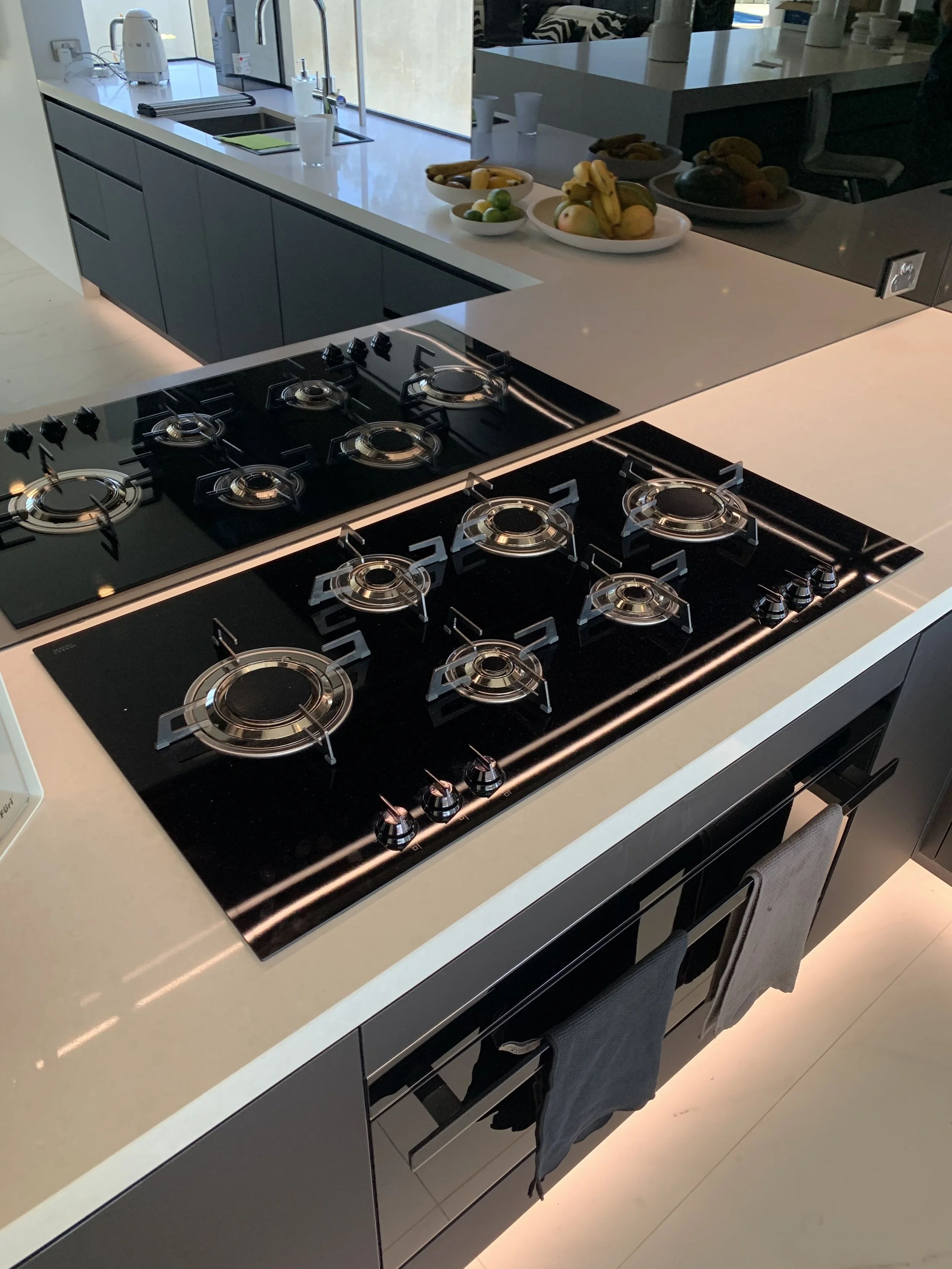 Modern kitchen with a black gas stove, white countertops, and bowls of fruit including bananas, apples, limes, and kiwis.