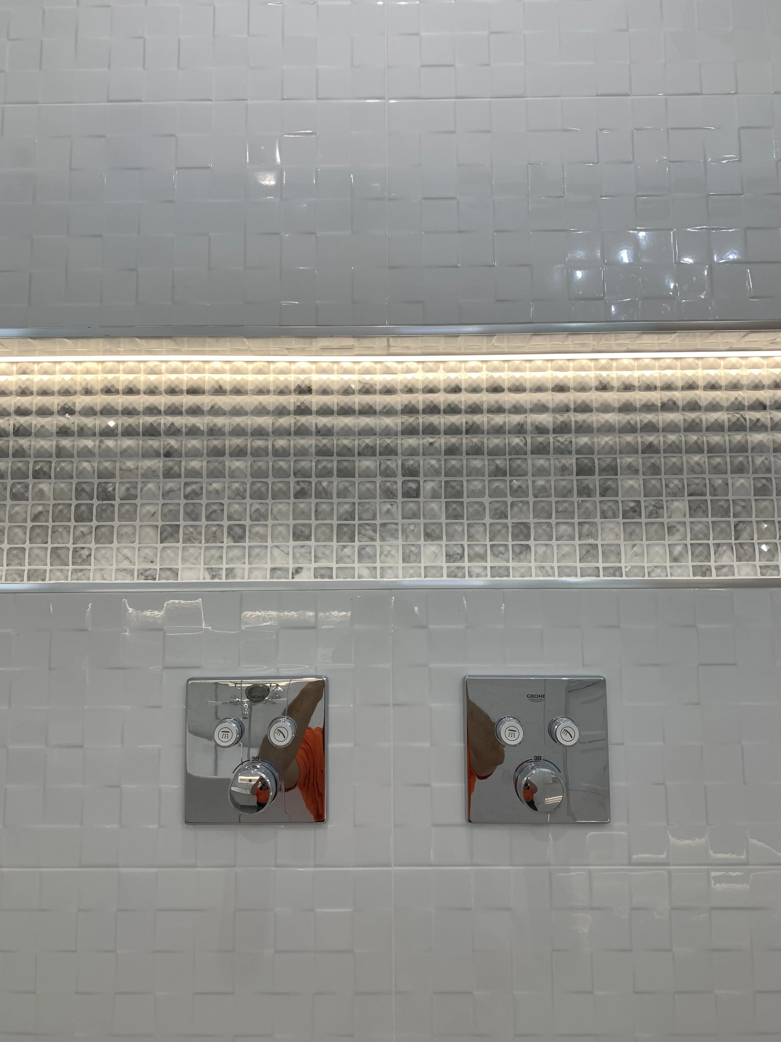 White tiled bathroom wall with metallic dual flush toilet buttons and decorative mosaic tile strip.