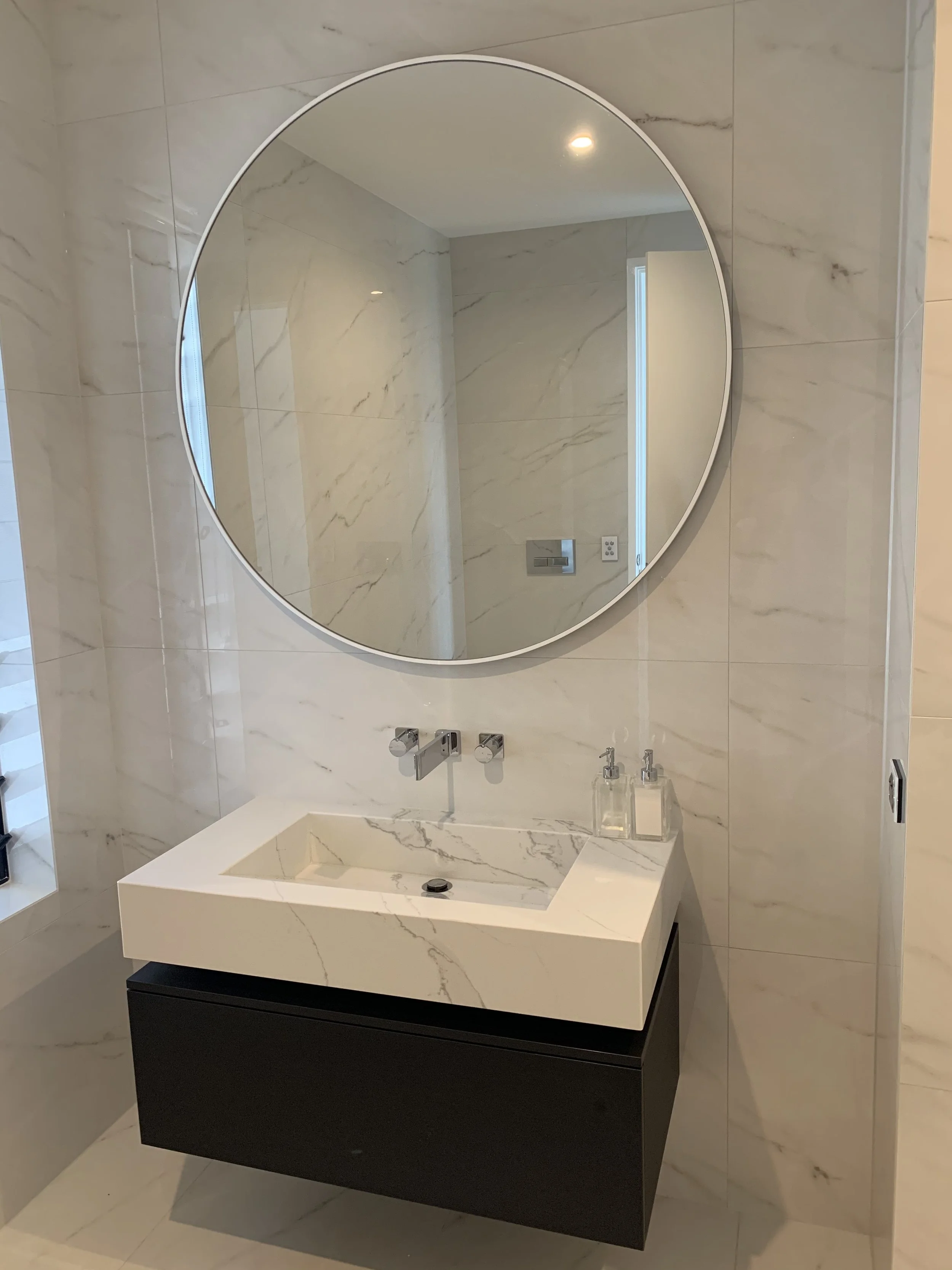 A modern bathroom vanity with a rectangular white marble sink, a round mirror above it, mounted on a marble wall, with a soap dispenser and a liquid soap bottle on the right side of the sink.