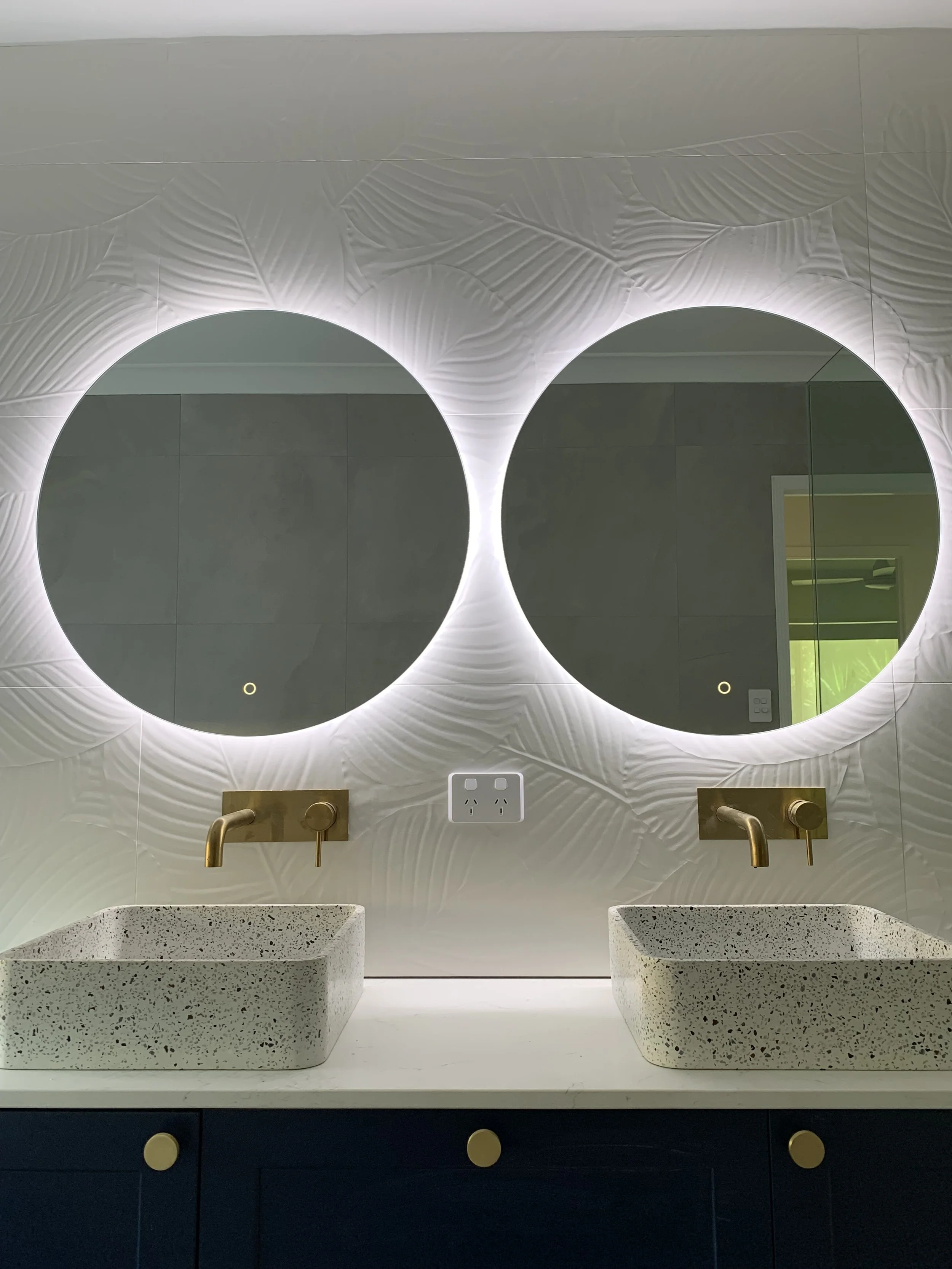 Modern bathroom with two round backlit mirrors, two rectangular terrazzo sinks, and gold faucets mounted on a textured wall.