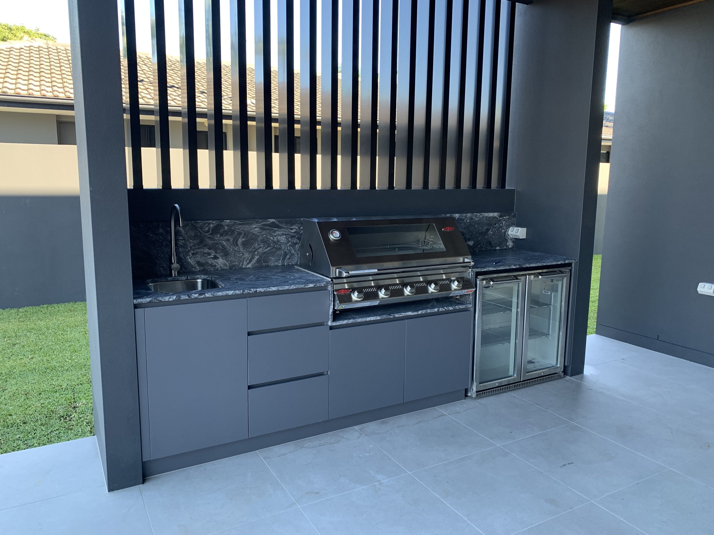 Outdoor kitchen area with a sink, a stainless steel grill, and a mini refrigerator, set against a black wall with vertical slats and a grey countertop.