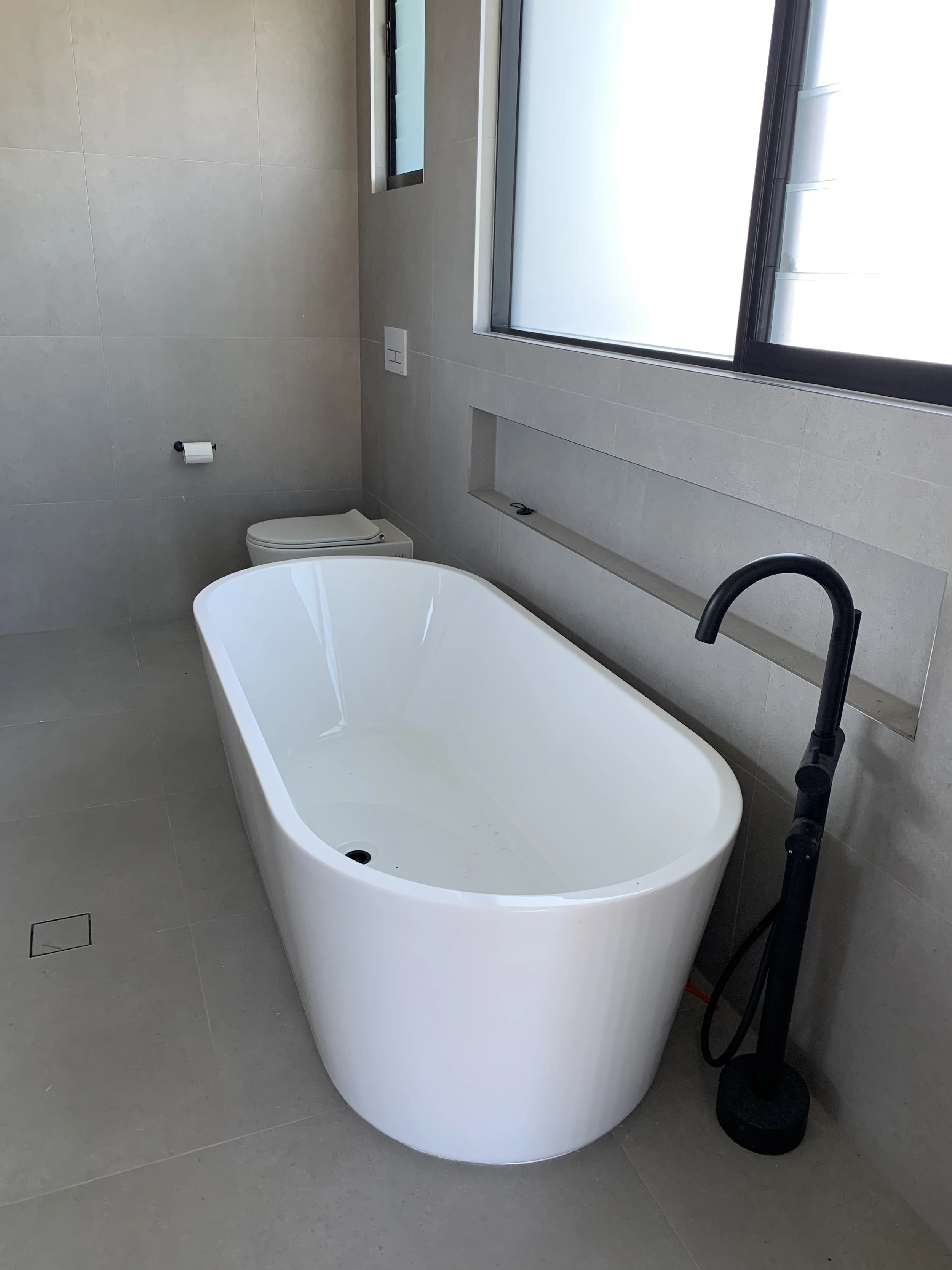 Modern bathroom with a white oval bathtub, black freestanding faucet, and a wall-mounted toilet, with large windows allowing natural light.