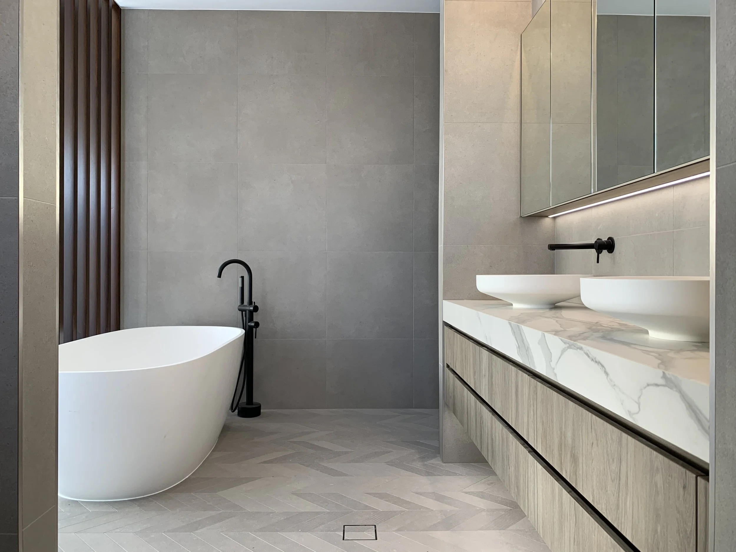 Modern bathroom with a free-standing bathtub, a dual sink vanity with a marble countertop, and large grey wall tiles.
