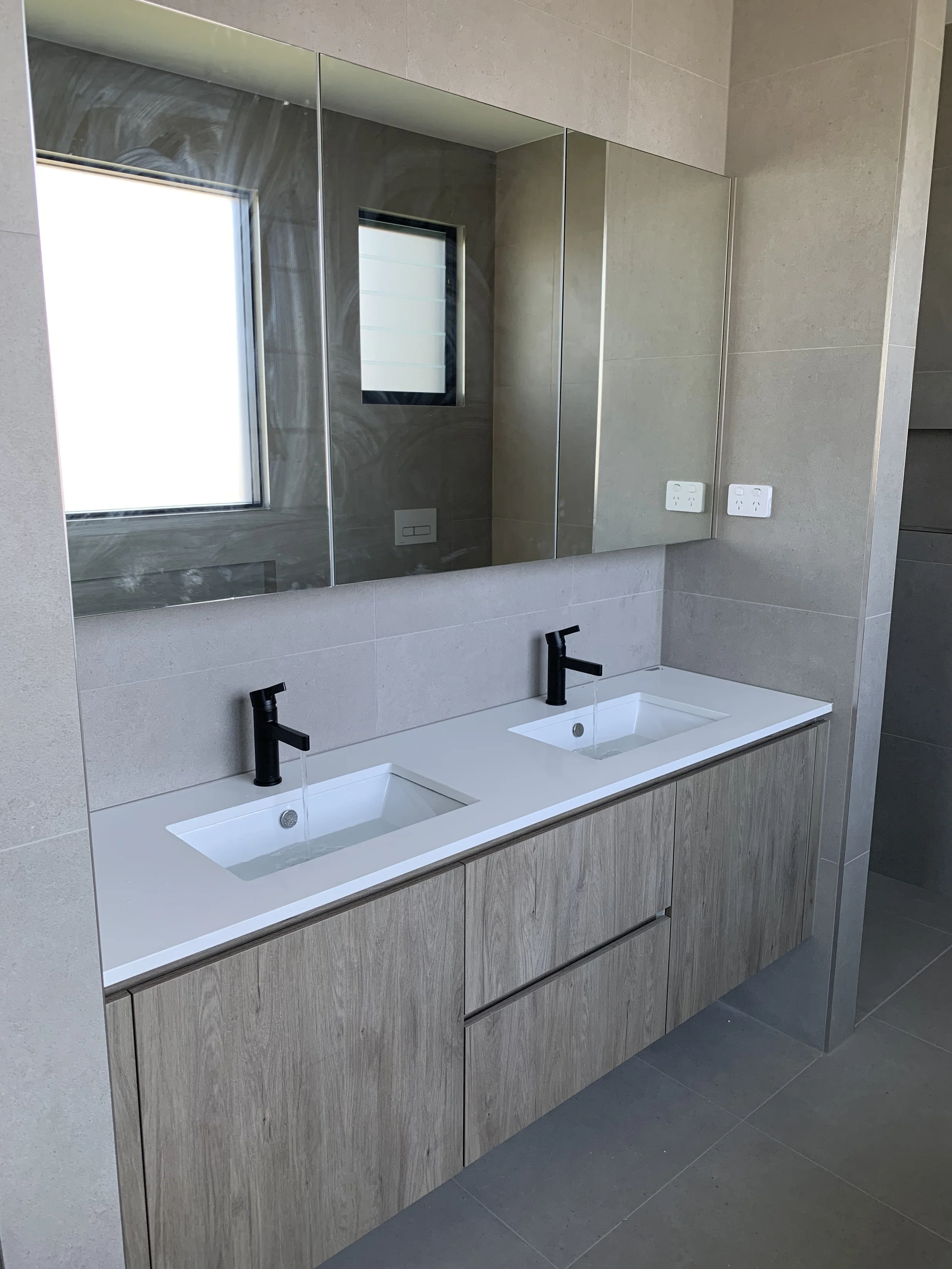 A modern bathroom with a double sink vanity featuring a white countertop, wood cabinets, and black fixtures. A large mirror with three panels is mounted above the sinks, reflecting a window that is also visible in the mirror. The bathroom has beige t