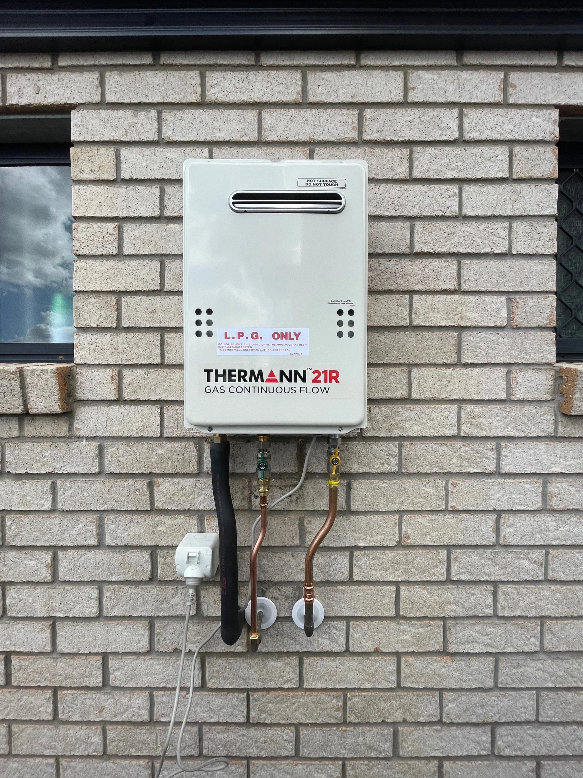 A gas tankless water heater mounted on a brick wall with pipes and a power cord connected to it.