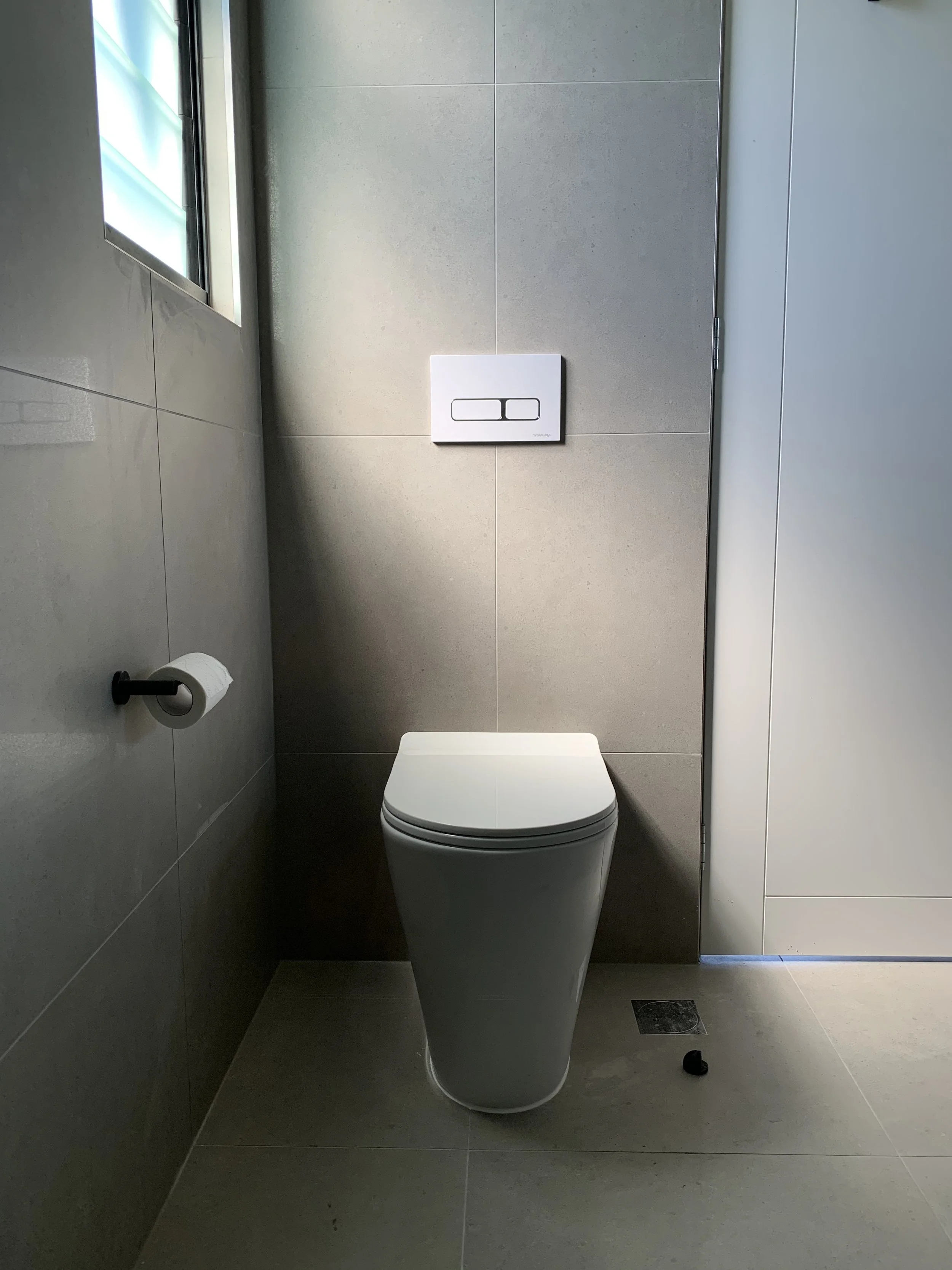 Modern bathroom with a wall-mounted toilet, toilet paper holder, window, and gray tiled walls and floor.