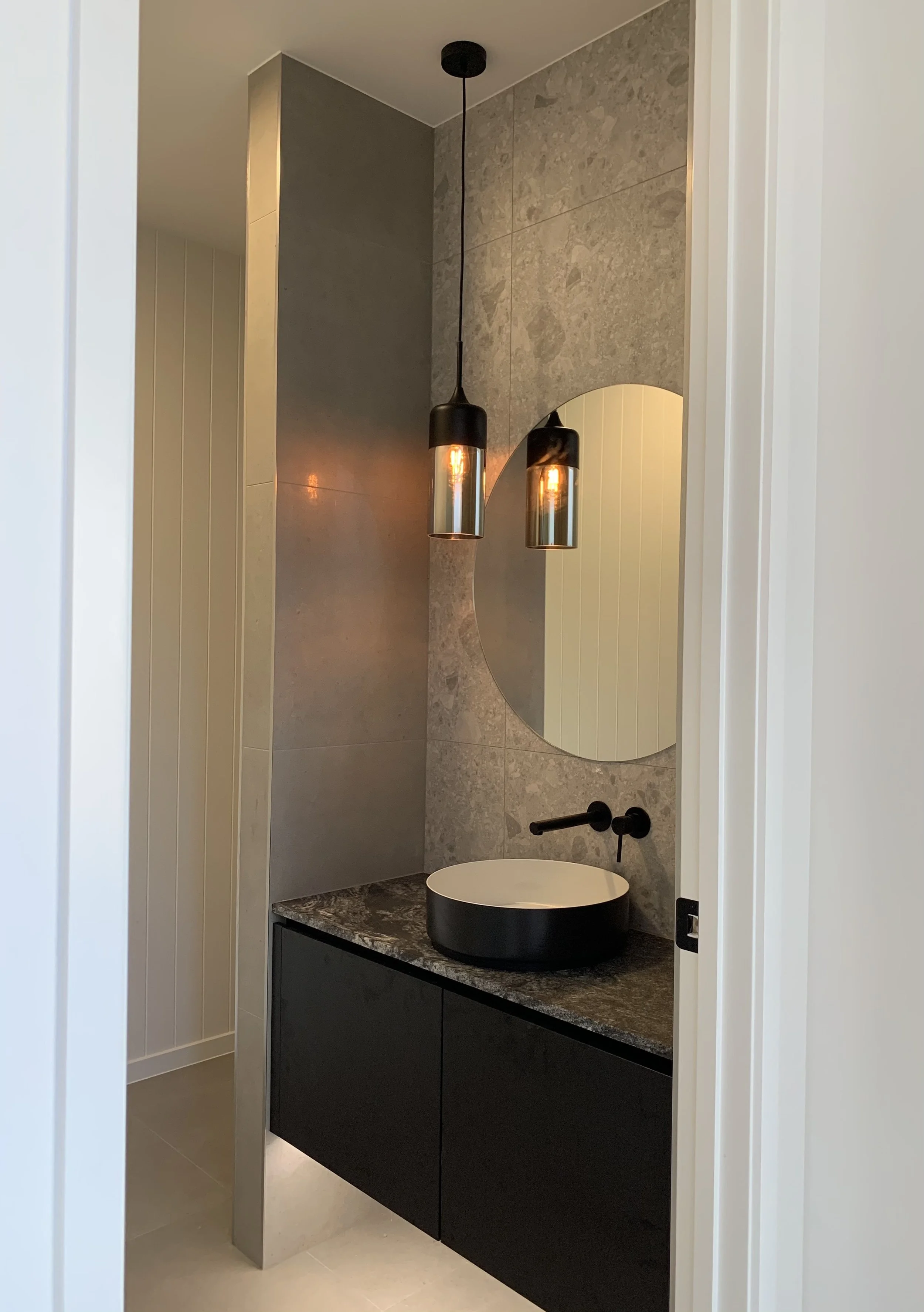 Modern bathroom vanity with a black circular vessel sink mounted on a dark marble countertop, a round mirror, and two black pendant lights hanging from the ceiling.