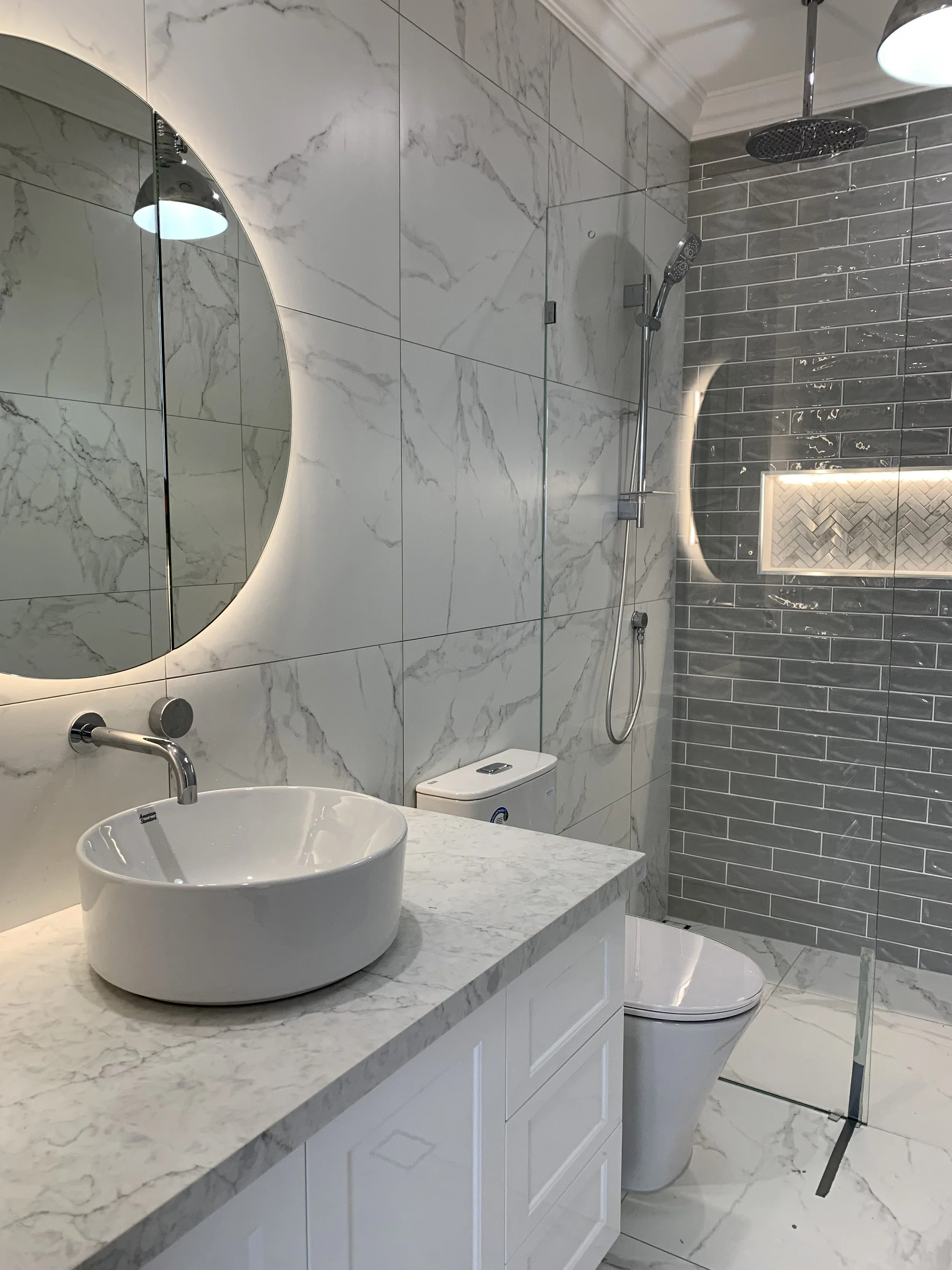 Modern bathroom with marble wall tiles, a round mirror with backlighting, a vessel sink on a marble countertop, a toilet, and a walk-in shower with grey subway tiles and a built-in niche, enclosed by a glass door.