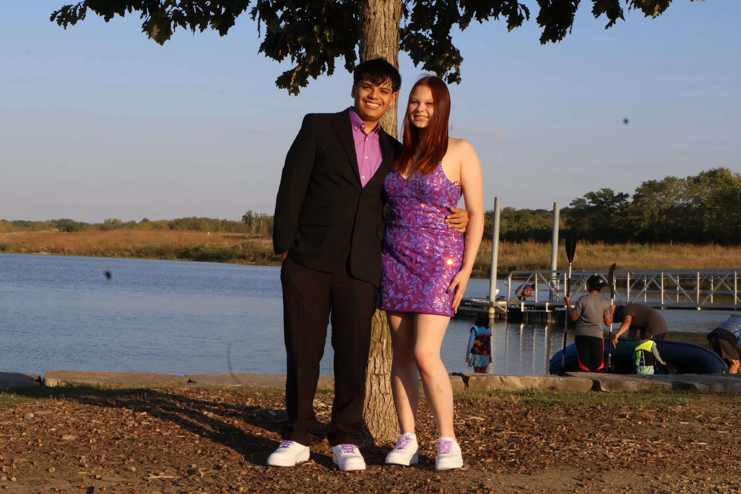 A young man and woman stand together by a tree near a lake, smiling. The man is wearing a black suit with a purple shirt, and the woman is in a purple patterned dress. There are people in the background at a dock and by the water, with a clear sky an