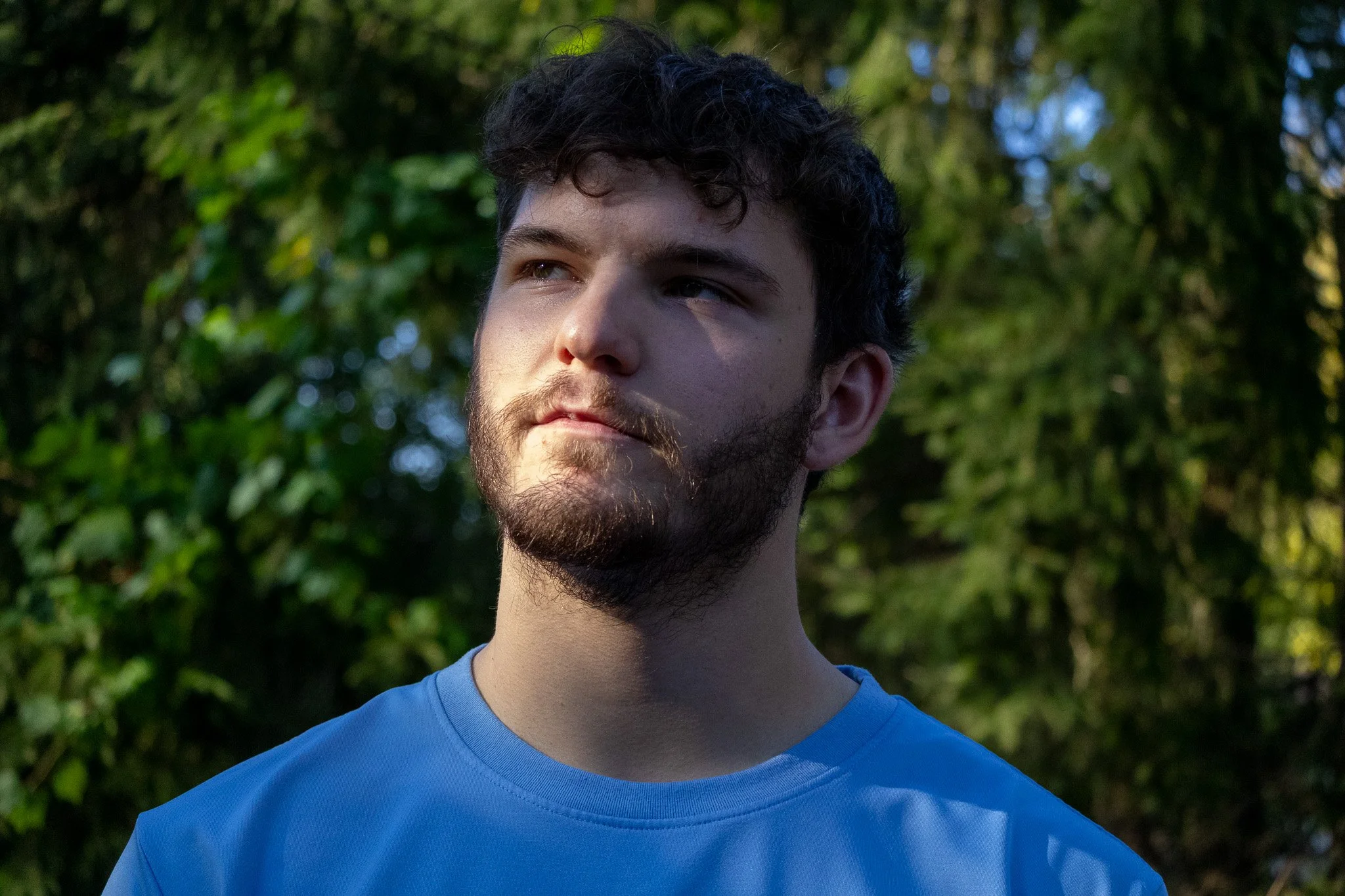 Young man with dark curly hair and a beard wearing a blue T-shirt outdoors in a wooded area with green foliage.