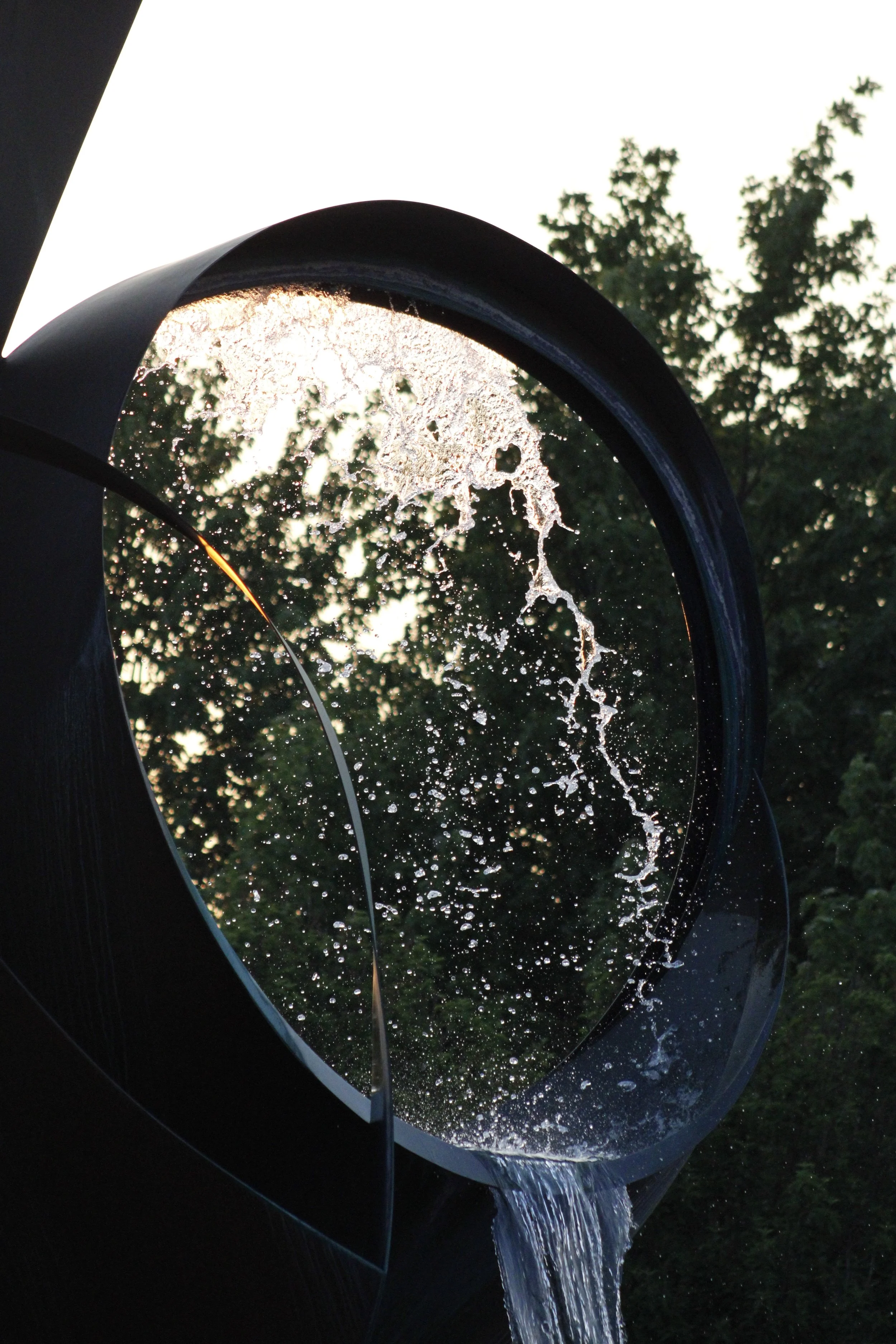Close-up of a modern outdoor water fountain with water flowing through a circular black metal structure, with trees and sky visible in the background.