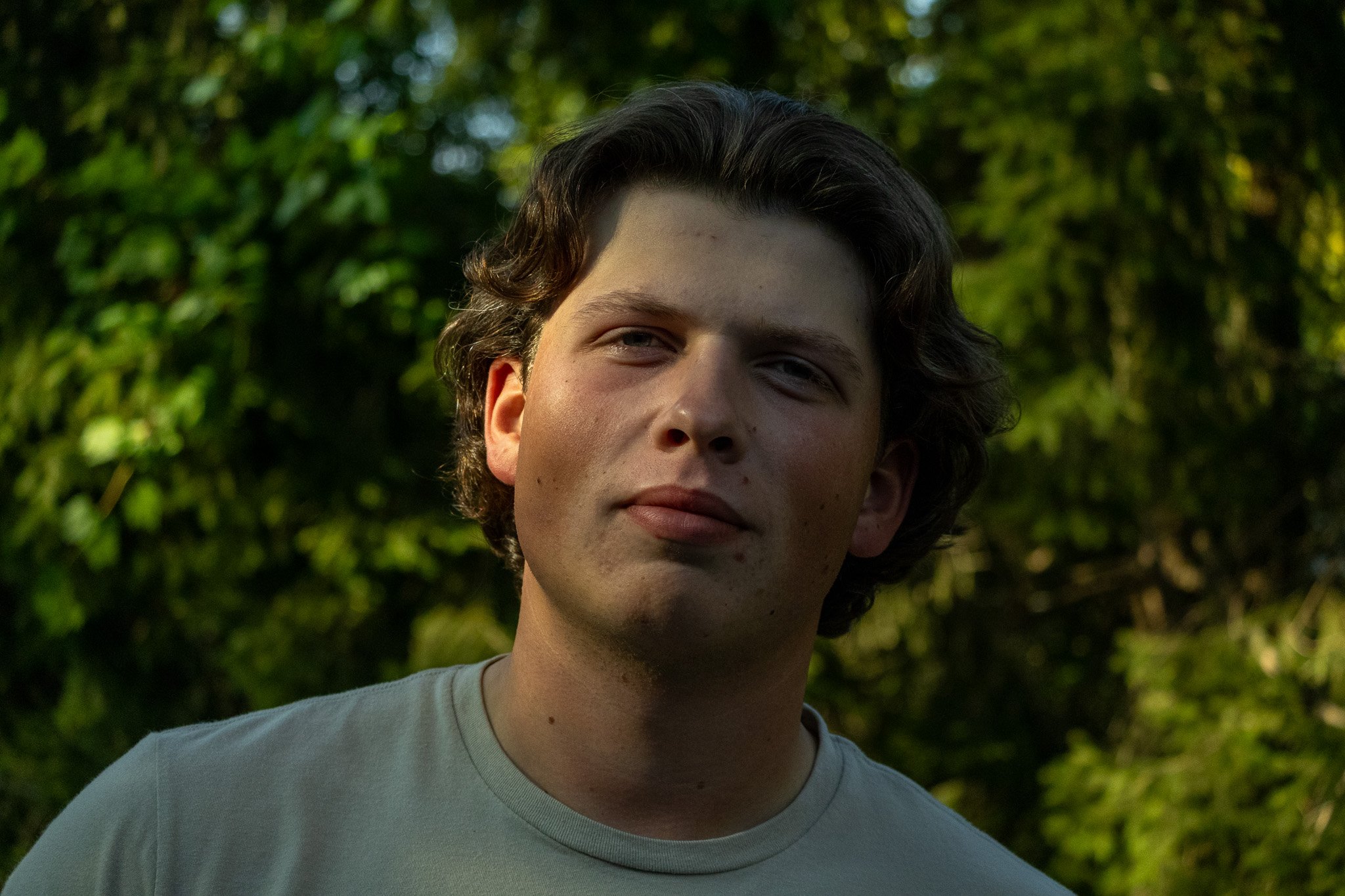 Young man with wavy brown hair and light skin outdoors in front of green trees, wearing a light gray t-shirt.