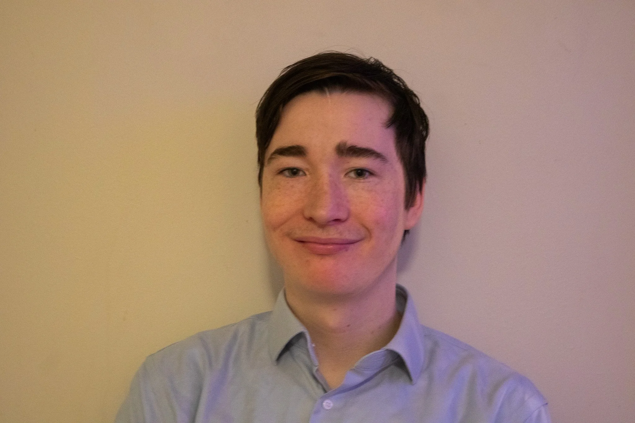 A young man with dark hair wearing a light-colored button-up shirt, smiling, standing against a plain wall.
