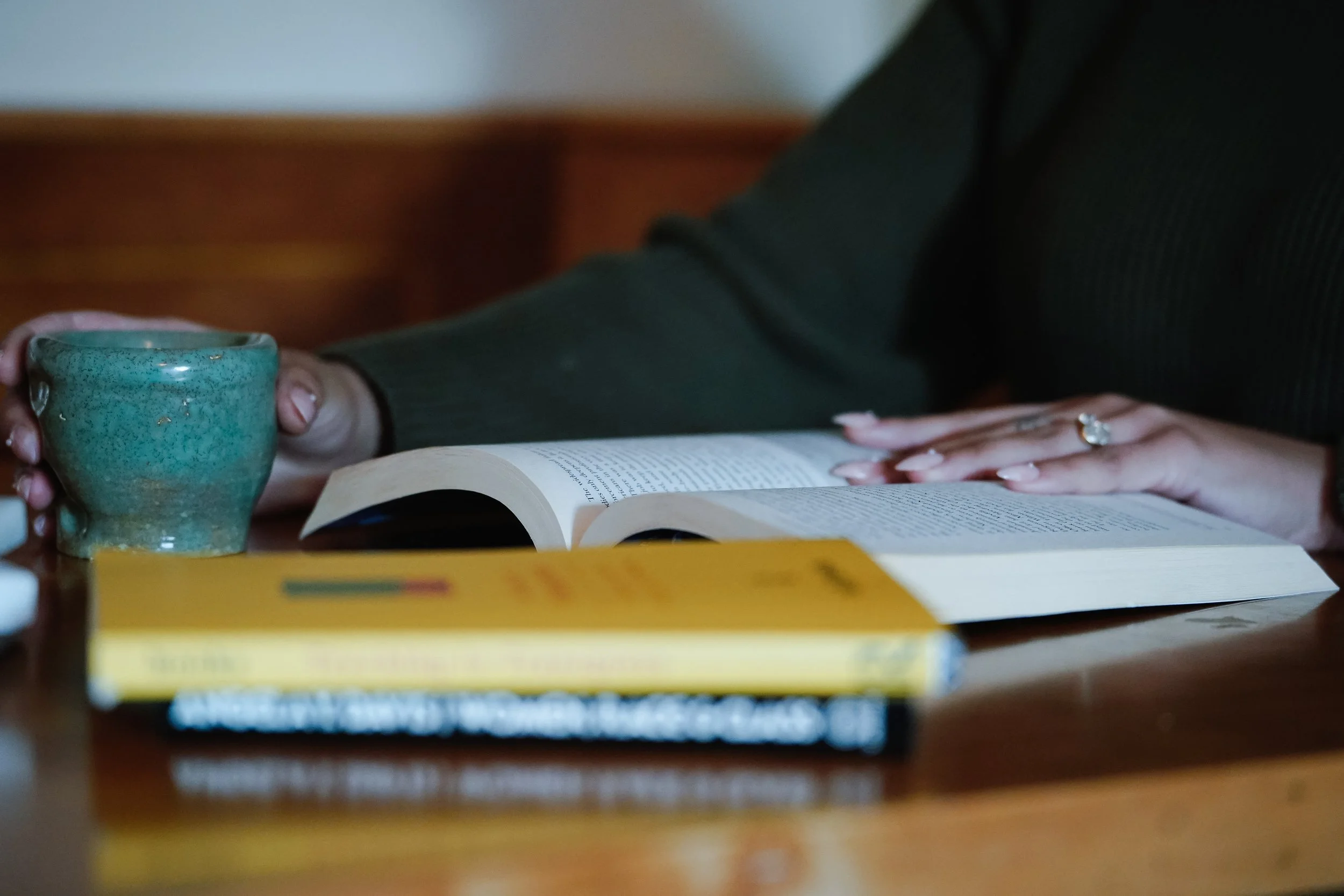 Person holding a blue ceramic mug while sitting at a table with an open book, a yellow book, and a stack of other books.