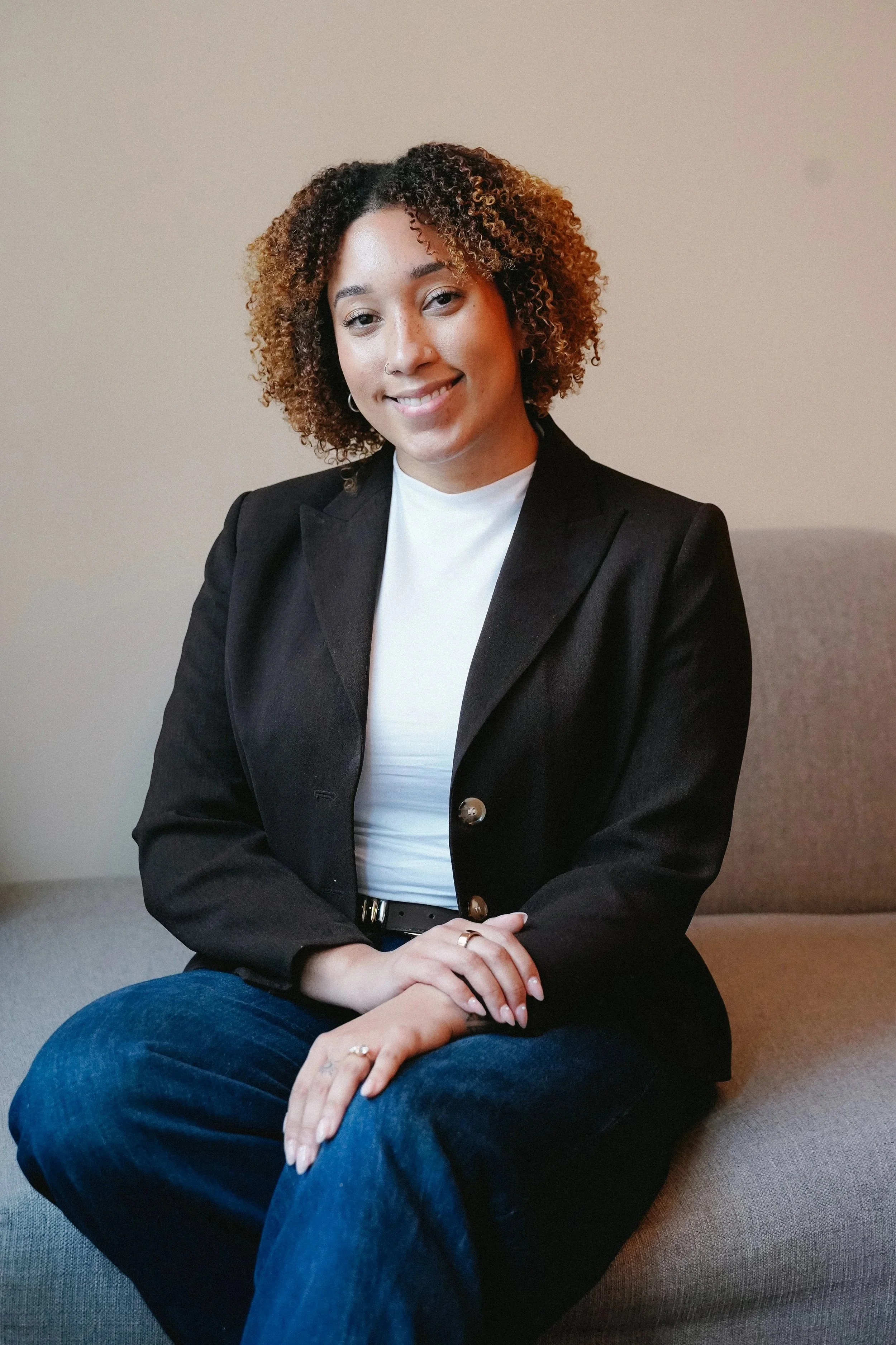 A Black woman with curly hair, wearing a black blazer over a white top, seated with her hands on her lap, smiling at the camera, against a beige background.