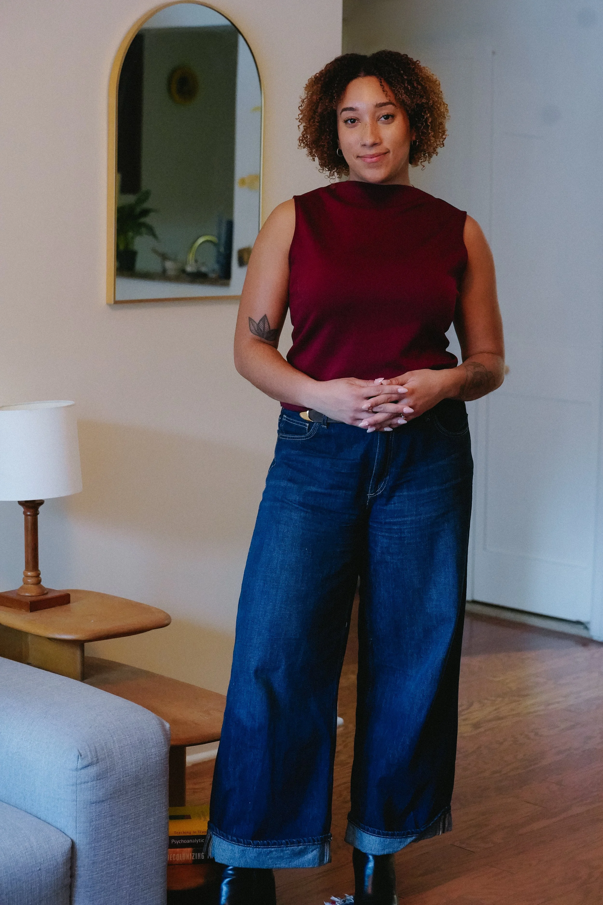 A woman with curly hair wearing a sleeveless maroon top and wide-leg blue jeans stands indoors near a mirror. A lamp and a book are on a side table next to her. The background includes a doorway and a glimpse of a kitchen.