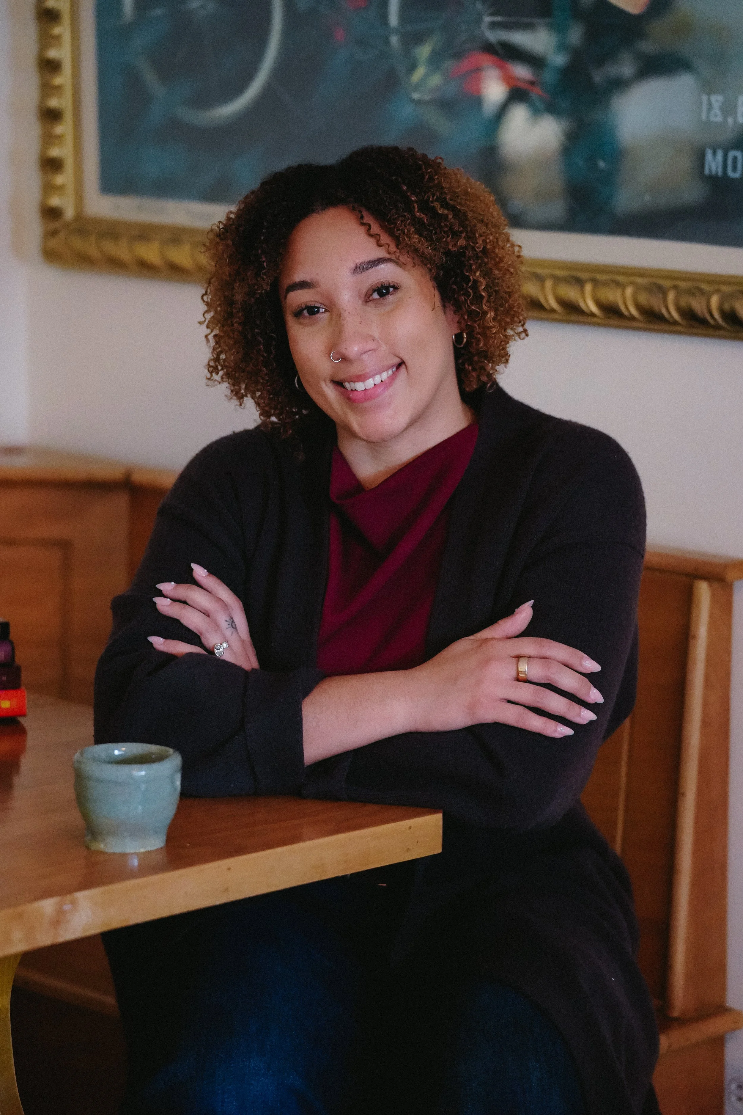 A woman with curly brown hair and a nose ring, sitting at a wooden table with arms crossed, smiling at the camera. A small ceramic mug is in front of her, and a framed artwork hangs on the wall behind her.