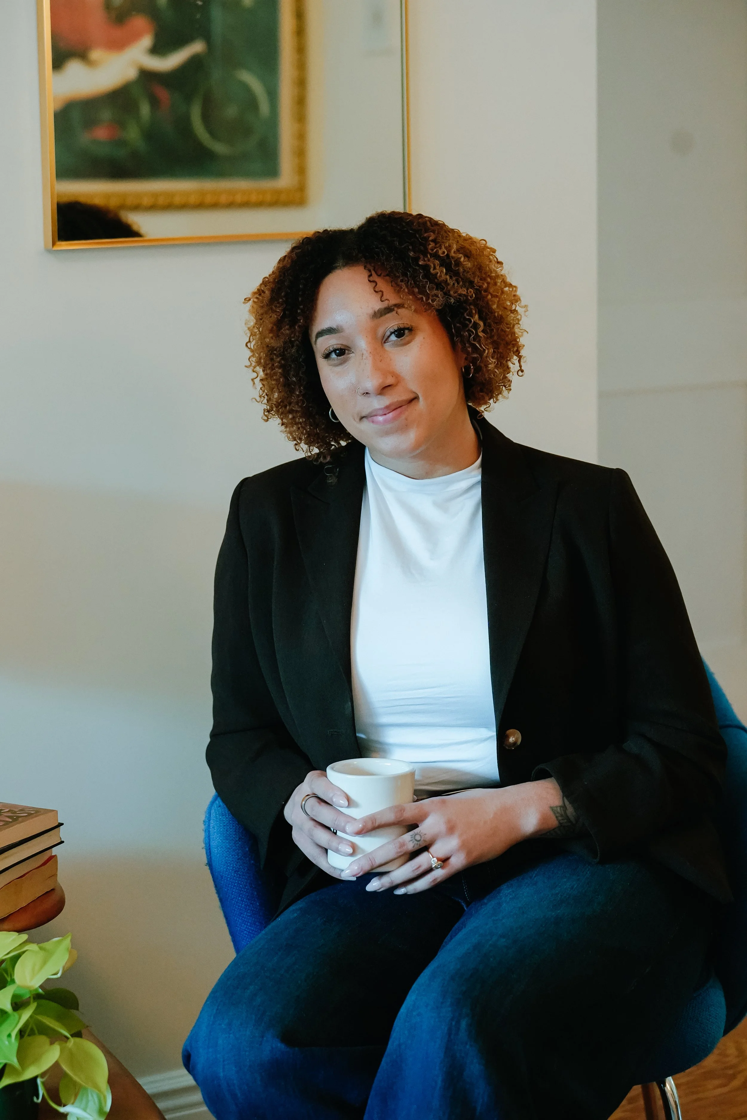 A woman sitting in a chair holding a white mug with both hands, wearing a black blazer over a white shirt, with short curly hair, in a room with a framed picture on the wall.