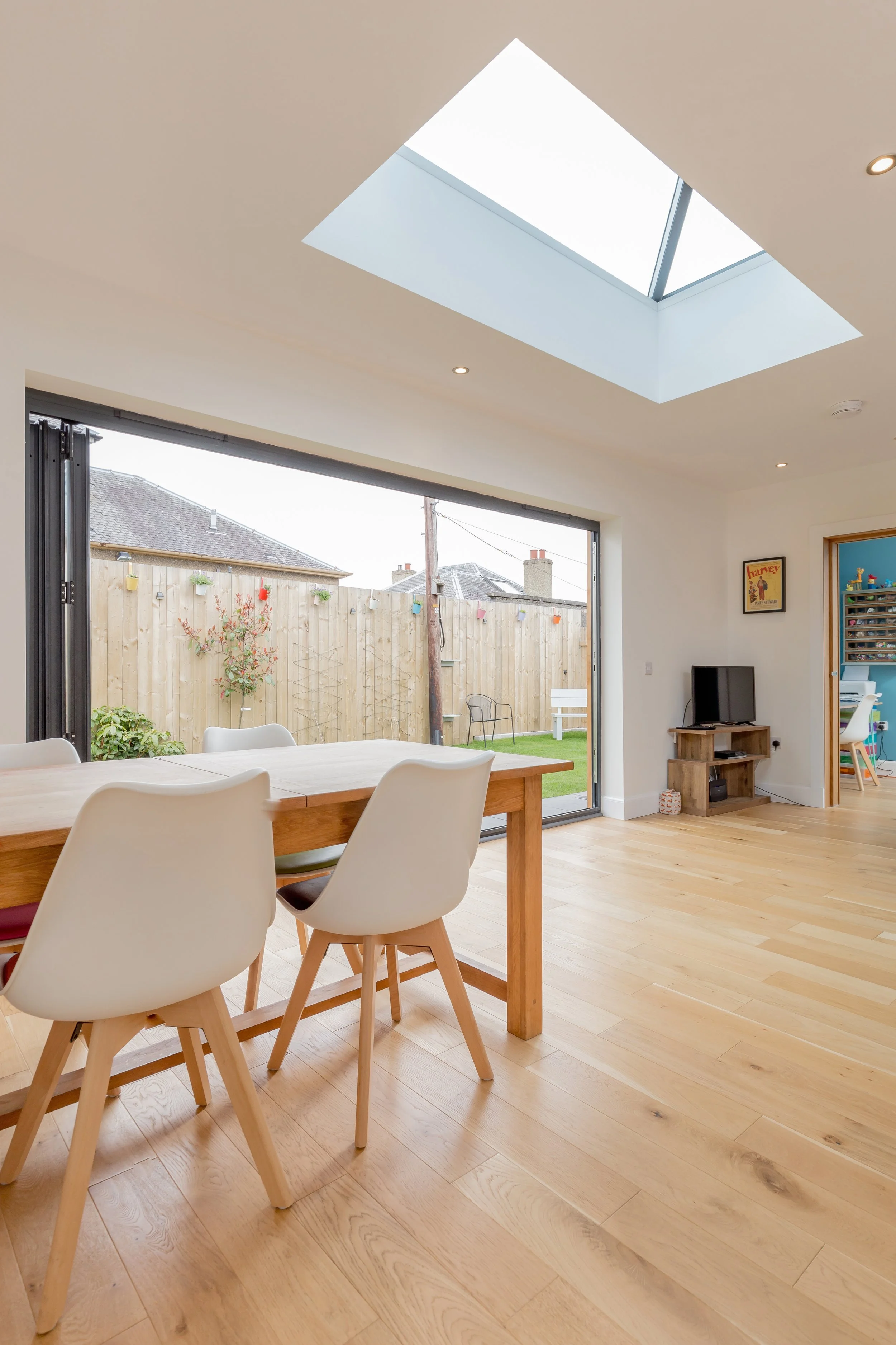 Interior view of a modern dining and living area with a skylight and sliding glass door opening to a backyard with a wooden fence.