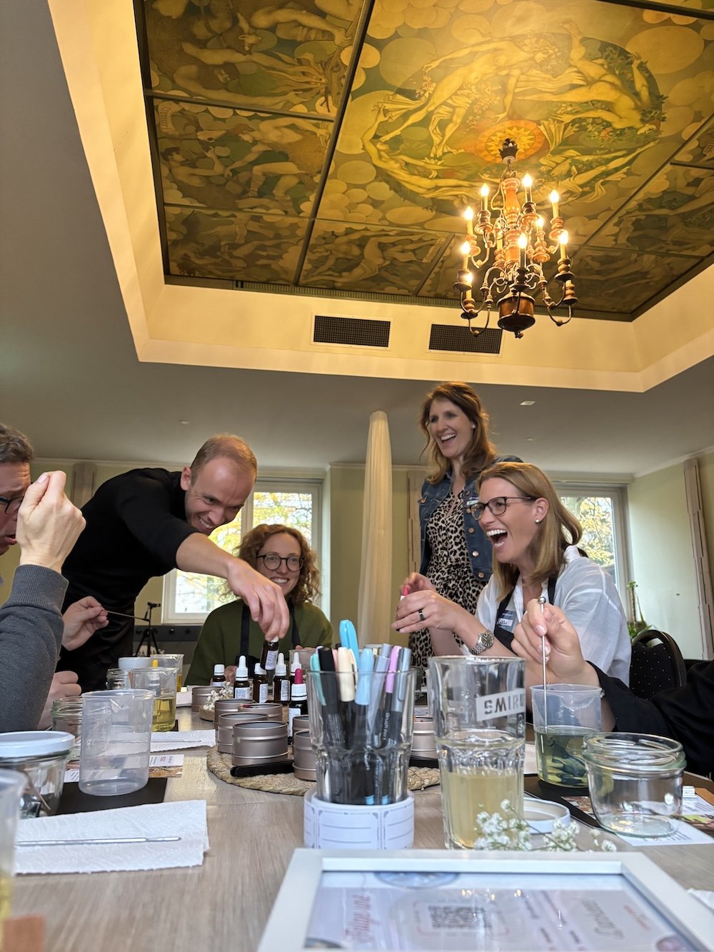 Menschen lachen und haben Spaß bei einem Kerzenworkshop. Sie sitzen gemeinmsam an einem Tisch in einem hellen Raum mit goldenen Deckenmalereien und einem Kronleuchter.