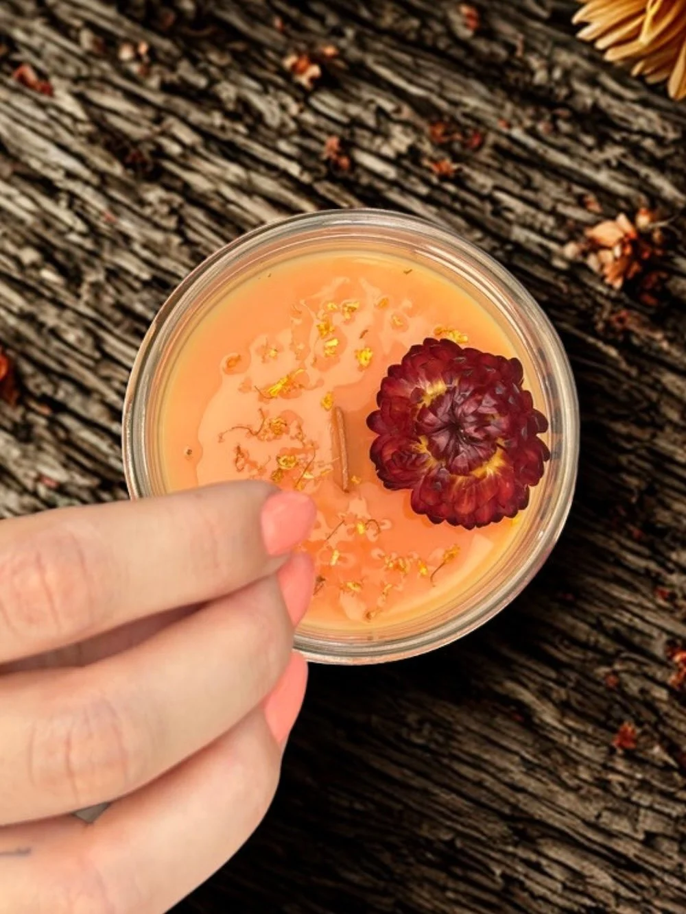 Nahaufnahme eines Glases mit orangefarbenem Wachs, verziert mit einer roten Blume und kleinen gelben Blüten, auf einem dunklen, holzigen Hintergrund.