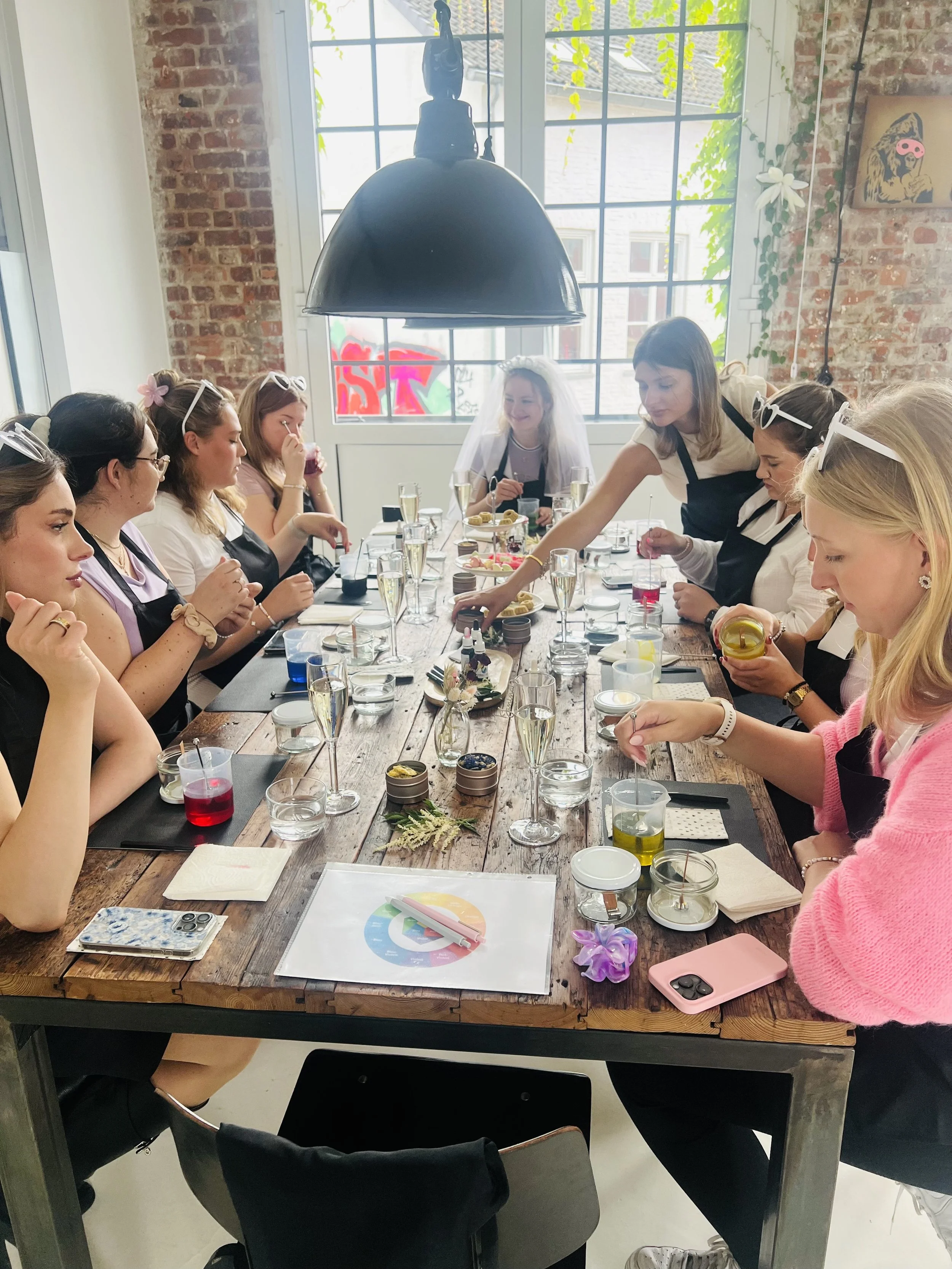Eine Gruppe von Frauen sitzt um einen Holztisch in einem Raum mit Backsteinwänden und großen Fenstern, bei einem JGA Kerzen-Workshop. Auf dem Tisch sind Getränke, kleine Tiegel mit Deko, Kerzen und Küchlein.