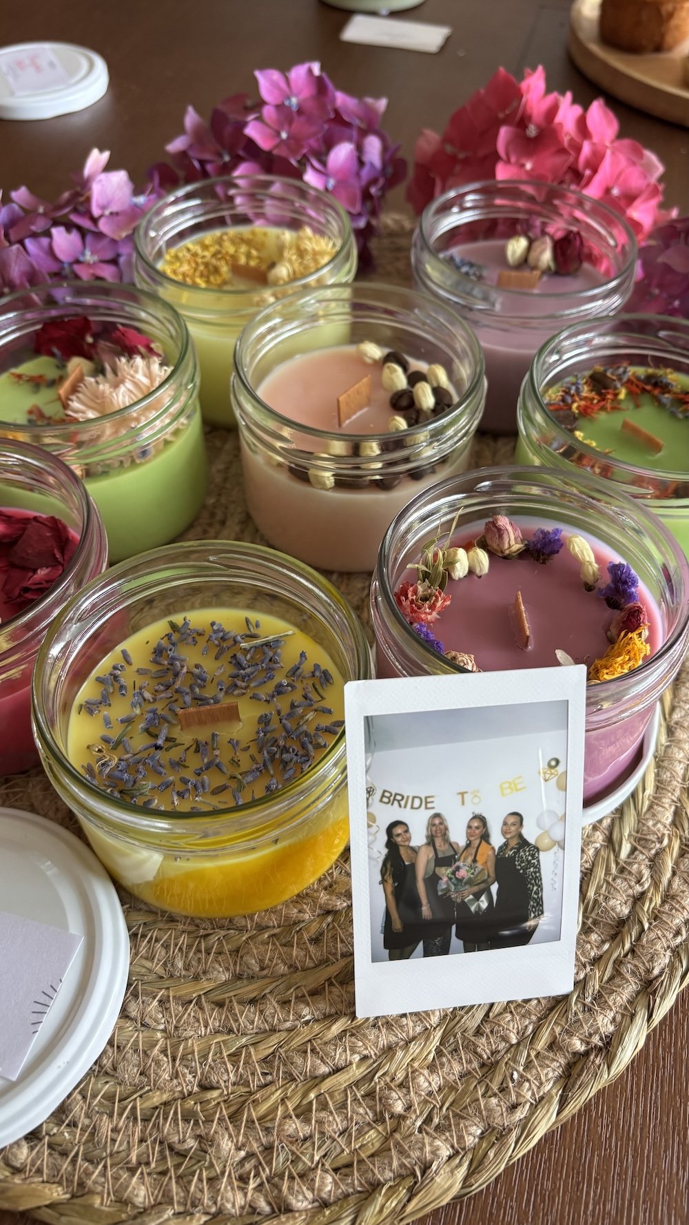 Verschiedene bunte Duftkerzen in Gläsern, Dekoration aus Blumen und eine kleine Polaroid-Foto einer Gruppe von Frauen, die einen JGA veranstalten.