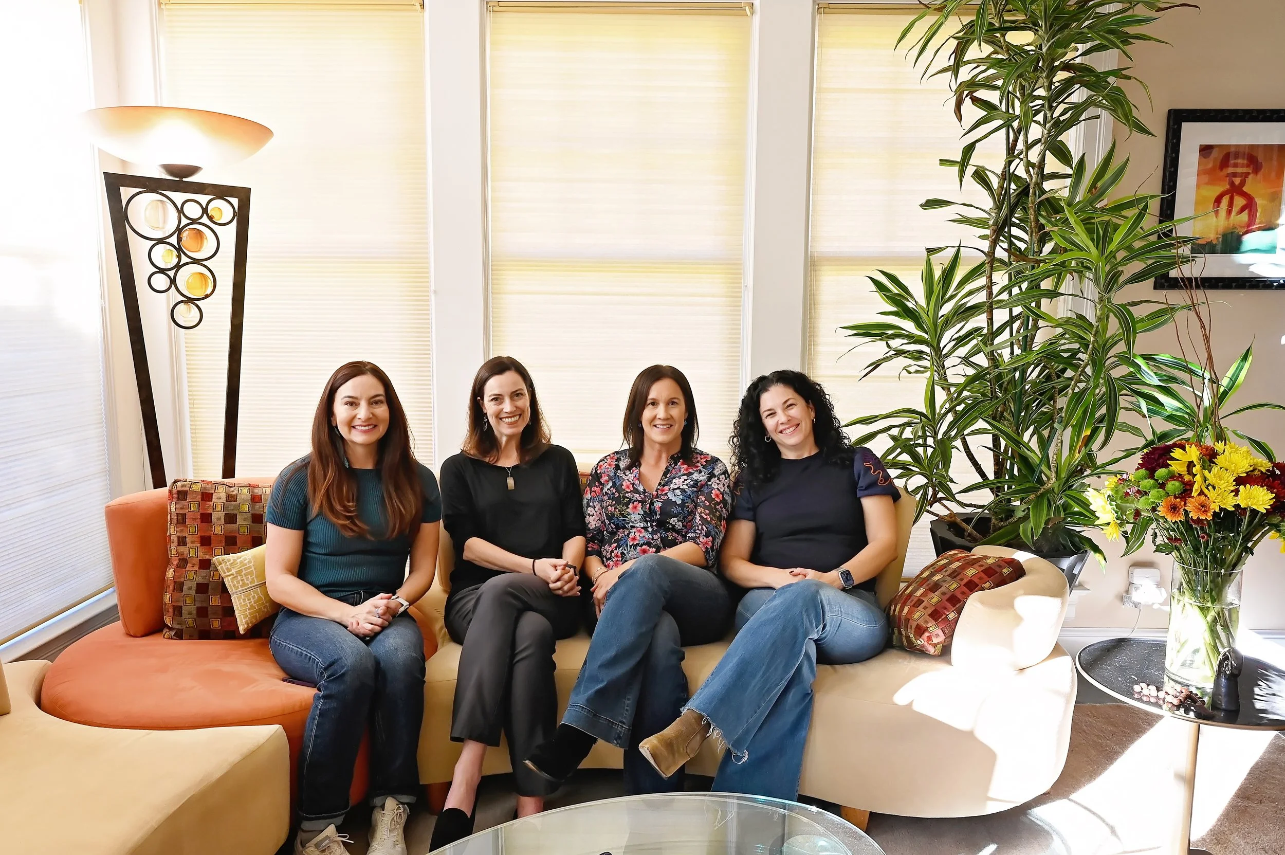 Four women sitting on a beige sectional sofa in a well-lit room with large window blinds, a tall decorative floor lamp, large green plants, a colorful flower arrangement on a side table, and a framed picture on the wall.