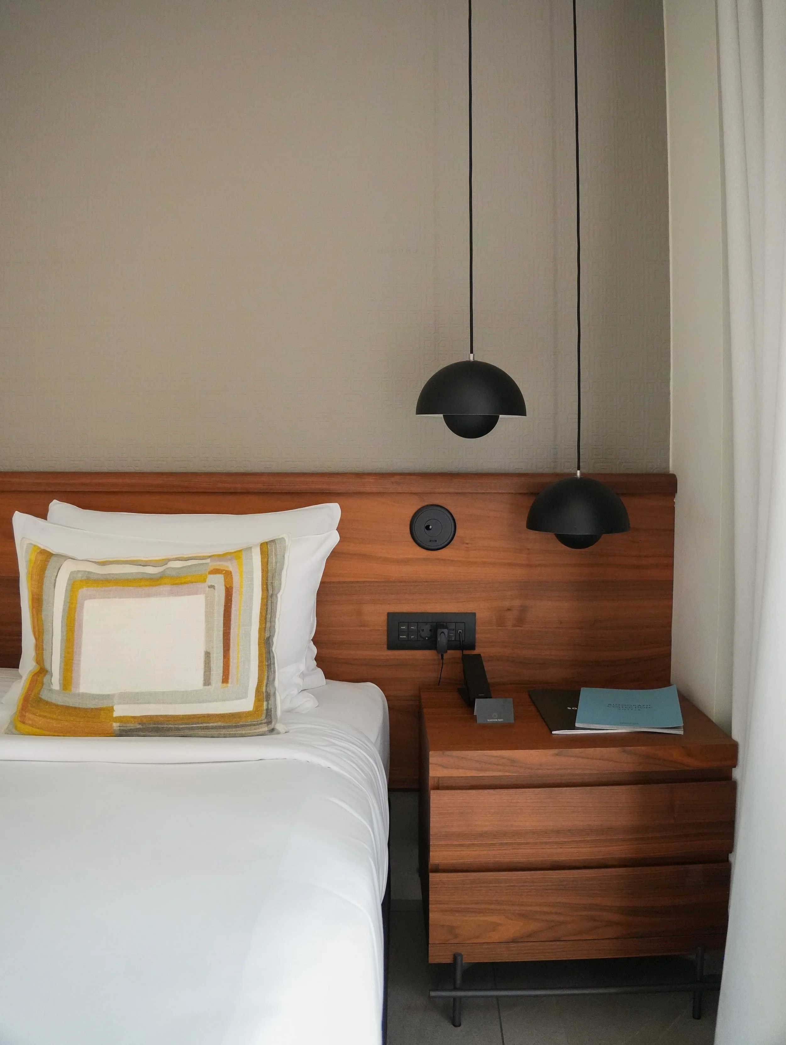 A bedroom with a white bed, a decorative pillow with geometric patterns, a wooden nightstand, hanging black pendant lights, and a wall with power outlets and a telephone.