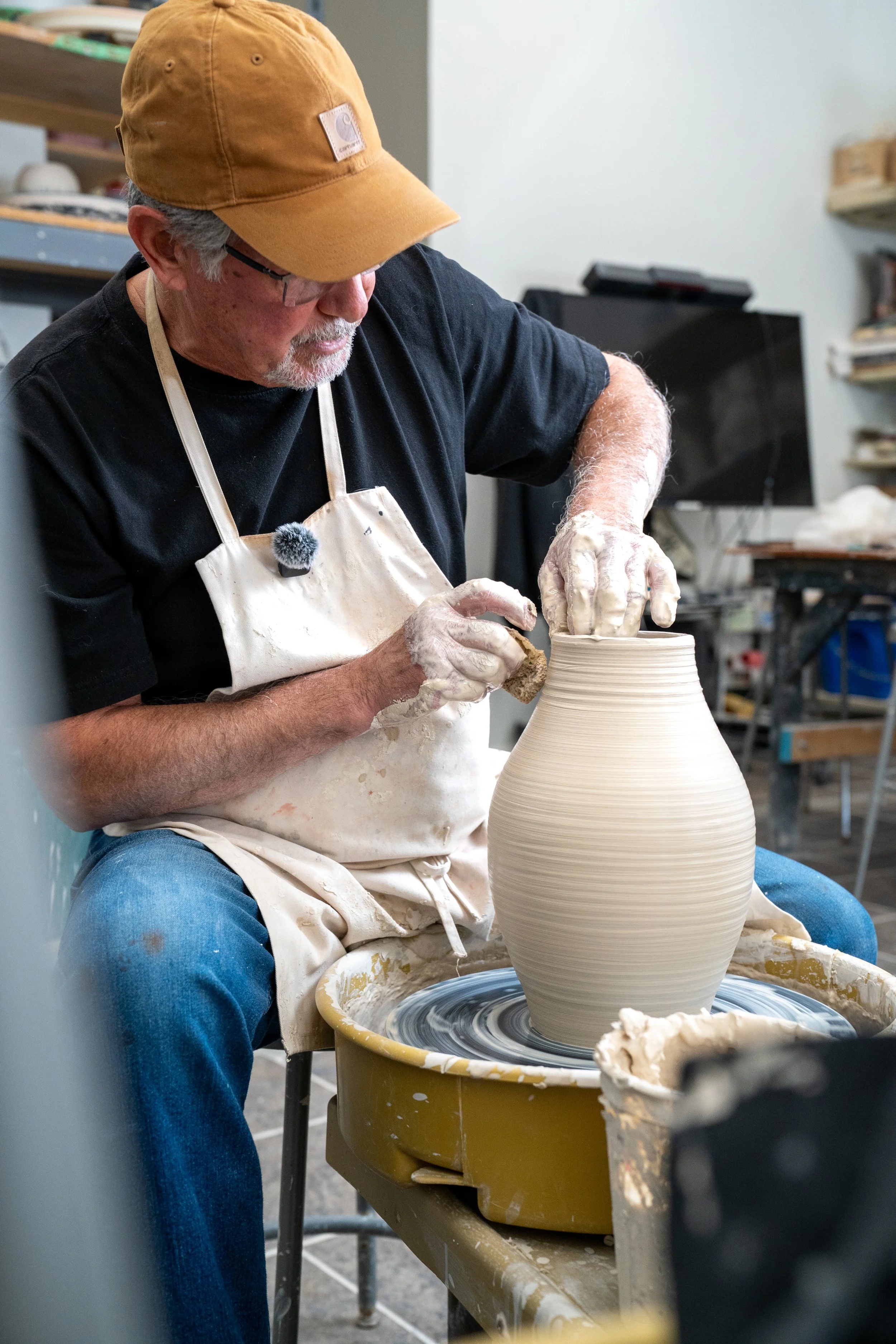 A man wearing a brown baseball cap, glasses, a black shirt, and a white apron is shaping a large, beige ceramic vase on a pottery wheel in a ceramics studio.