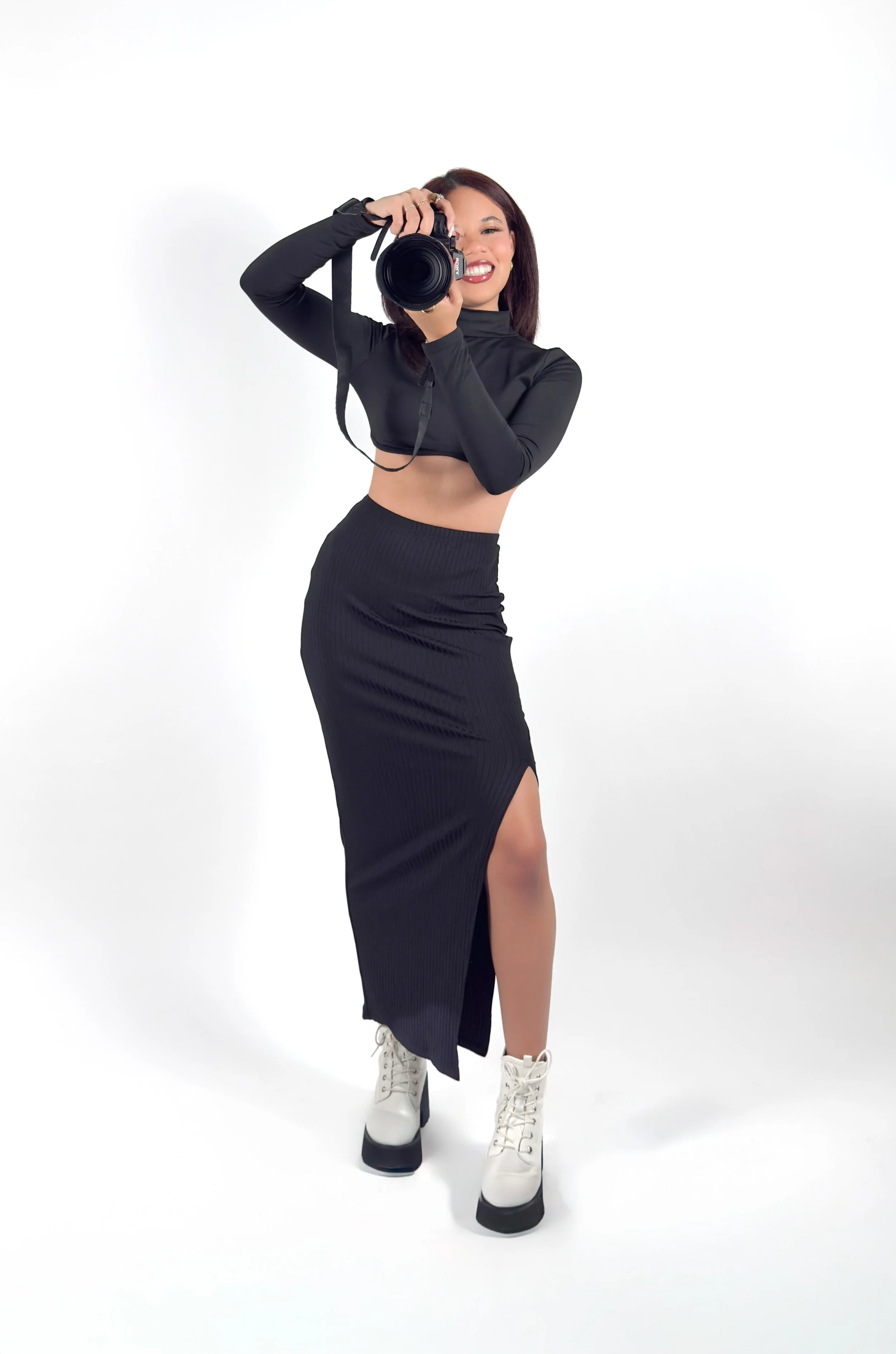 Woman dressed in black, holding a camera and smiling in a studio with a white background.