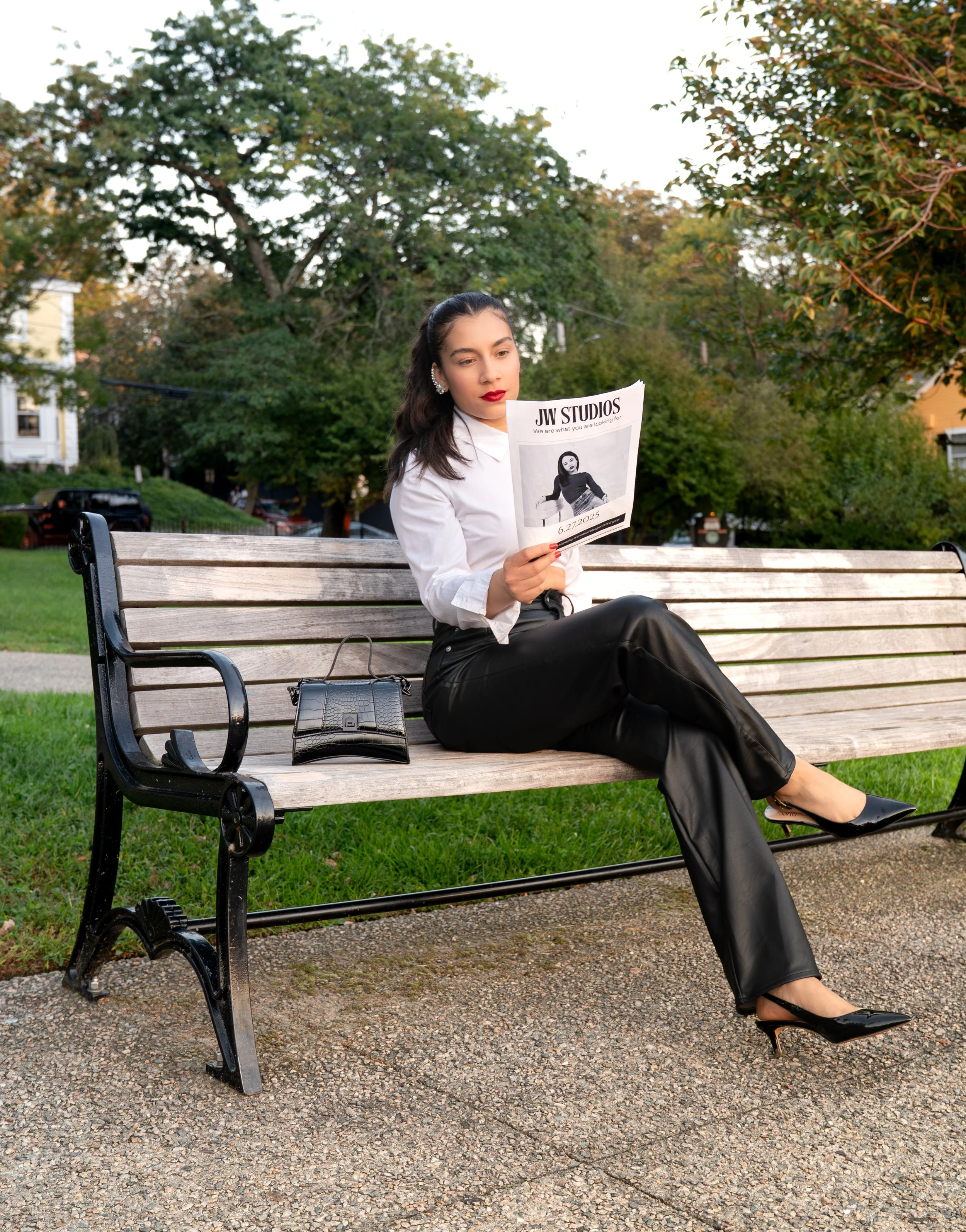 A woman sitting on a park bench reading a newspaper titled 'JW STUDIOS' with a black handbag beside her, outdoors in a green park with trees and houses in the background.