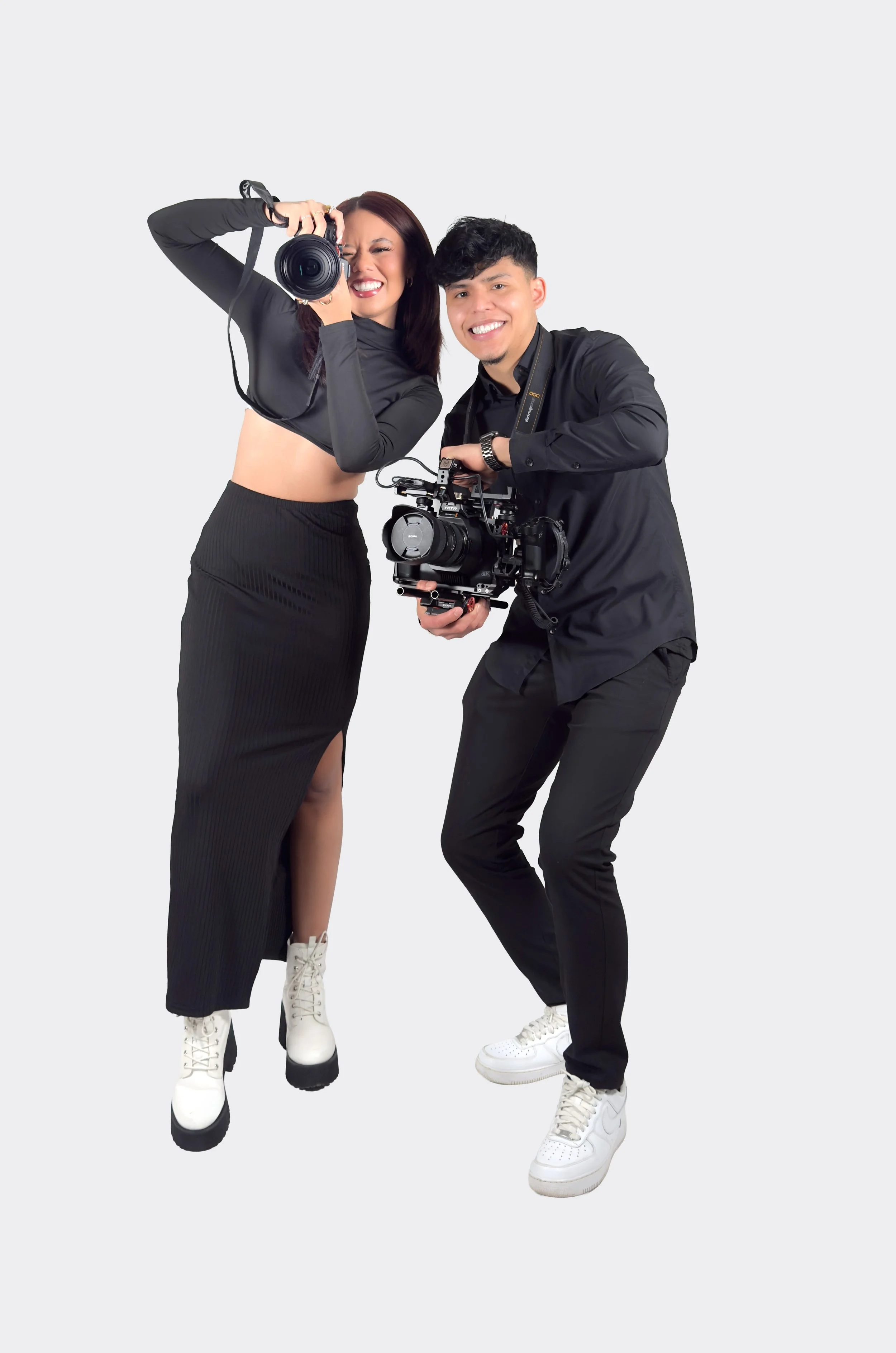 Two people, a woman and a man, holding professional cameras, posing and smiling for a photo against a plain background.