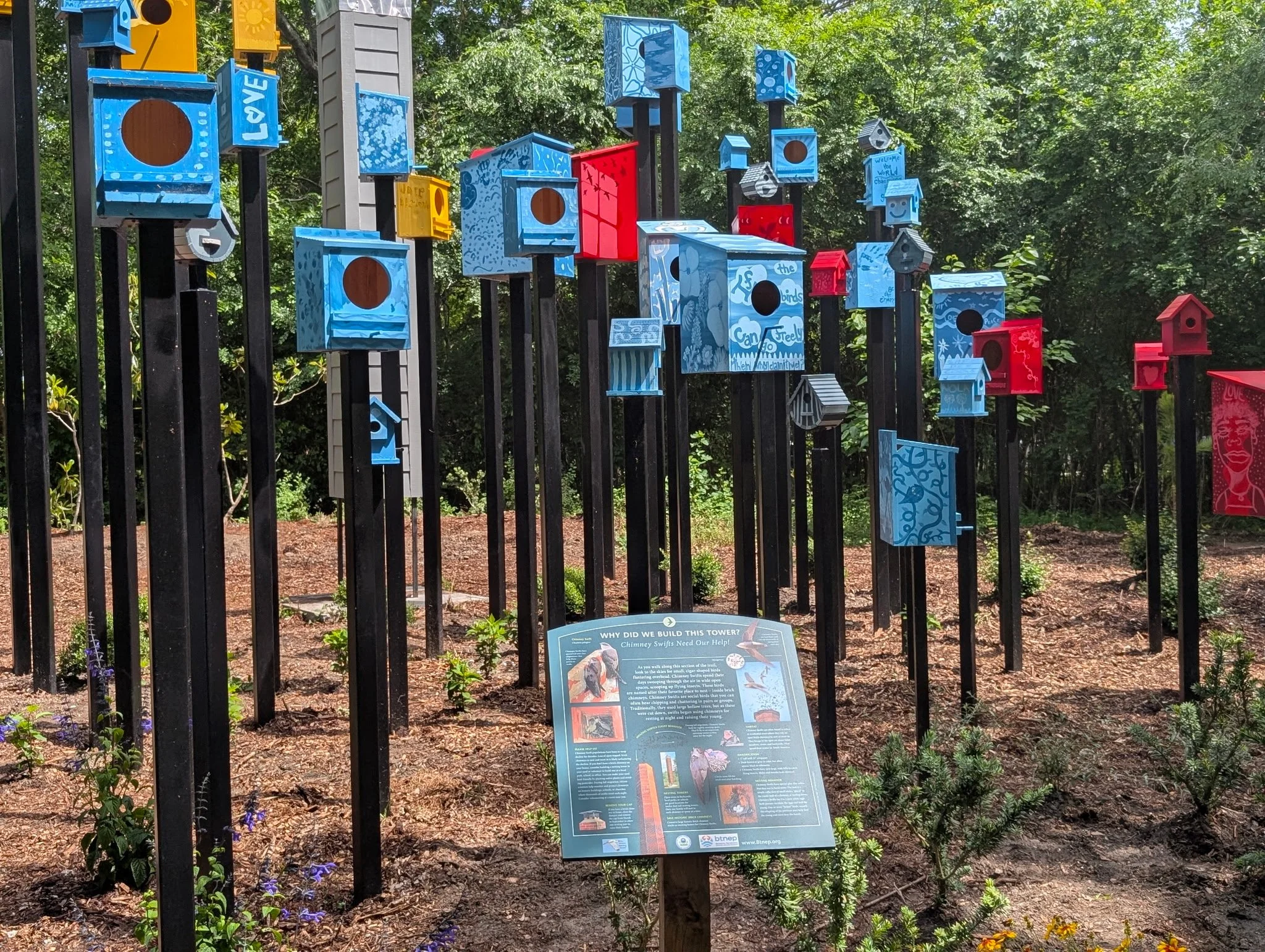 A display of colorful birdhouses mounted on black poles outdoors, with a sign explaining the significance of chimney swifts and their nesting habits.