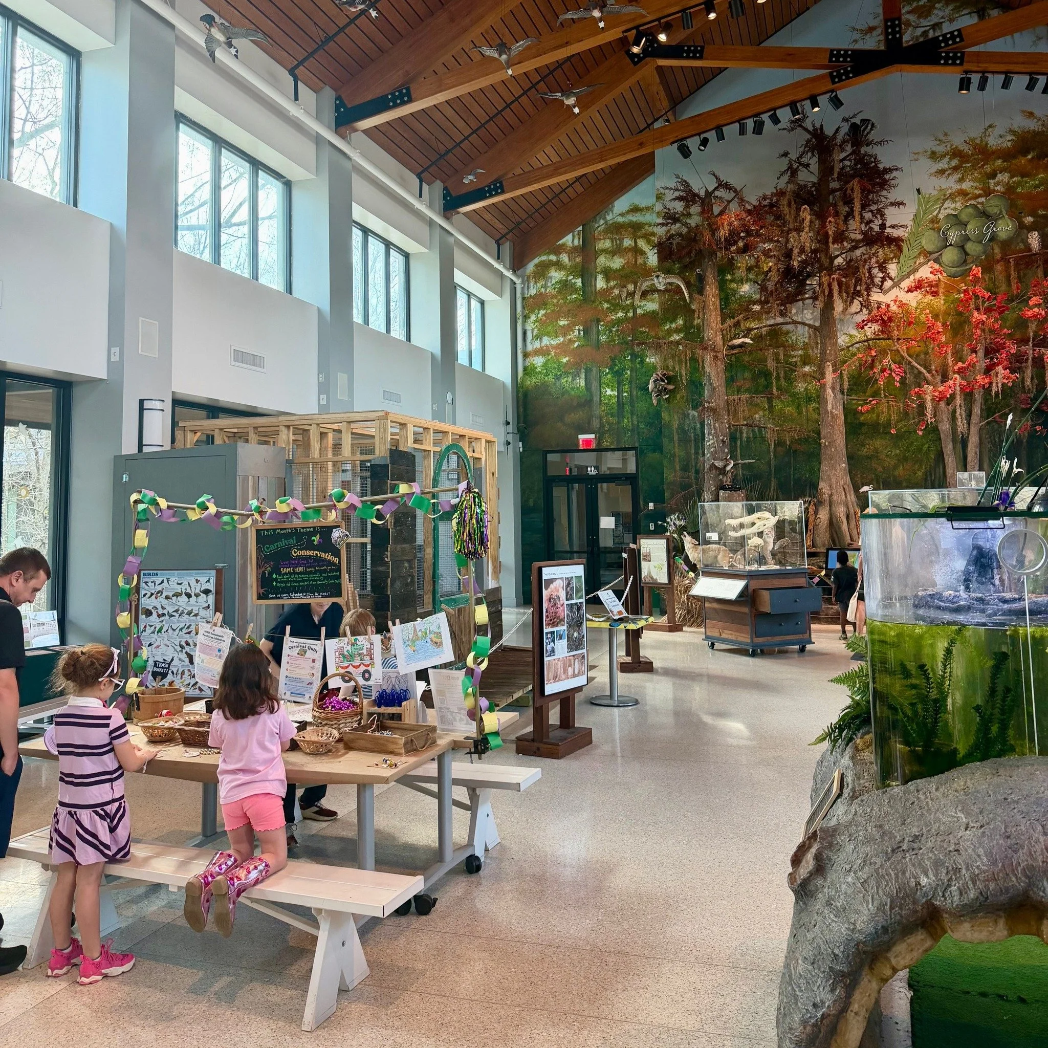 Children gather around tables at an indoor educational exhibit, with a forest mural and a rock formation in the background.