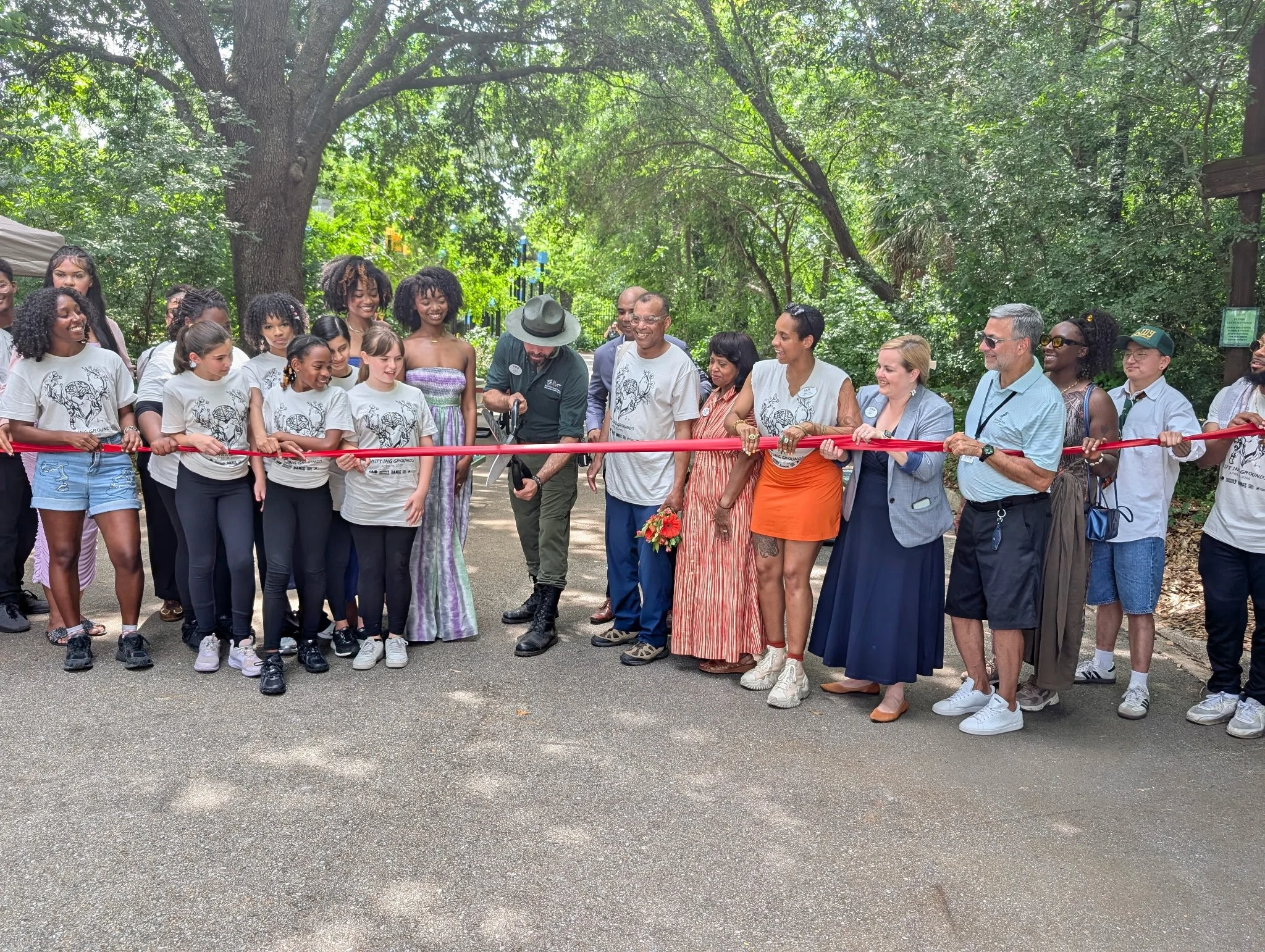 A diverse group of people gathered outdoors for a ribbon-cutting ceremony in a park with tall trees. The group includes children, adults, and seniors, with some smiling and looking at the ribbon being cut.