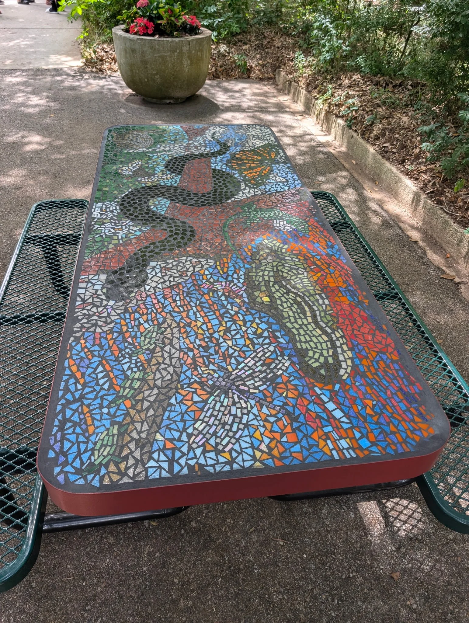 Outdoor table with a colorful mosaic snake design, surrounded by green metal chairs, on a concrete surface with a large flower pot and foliage in the background.